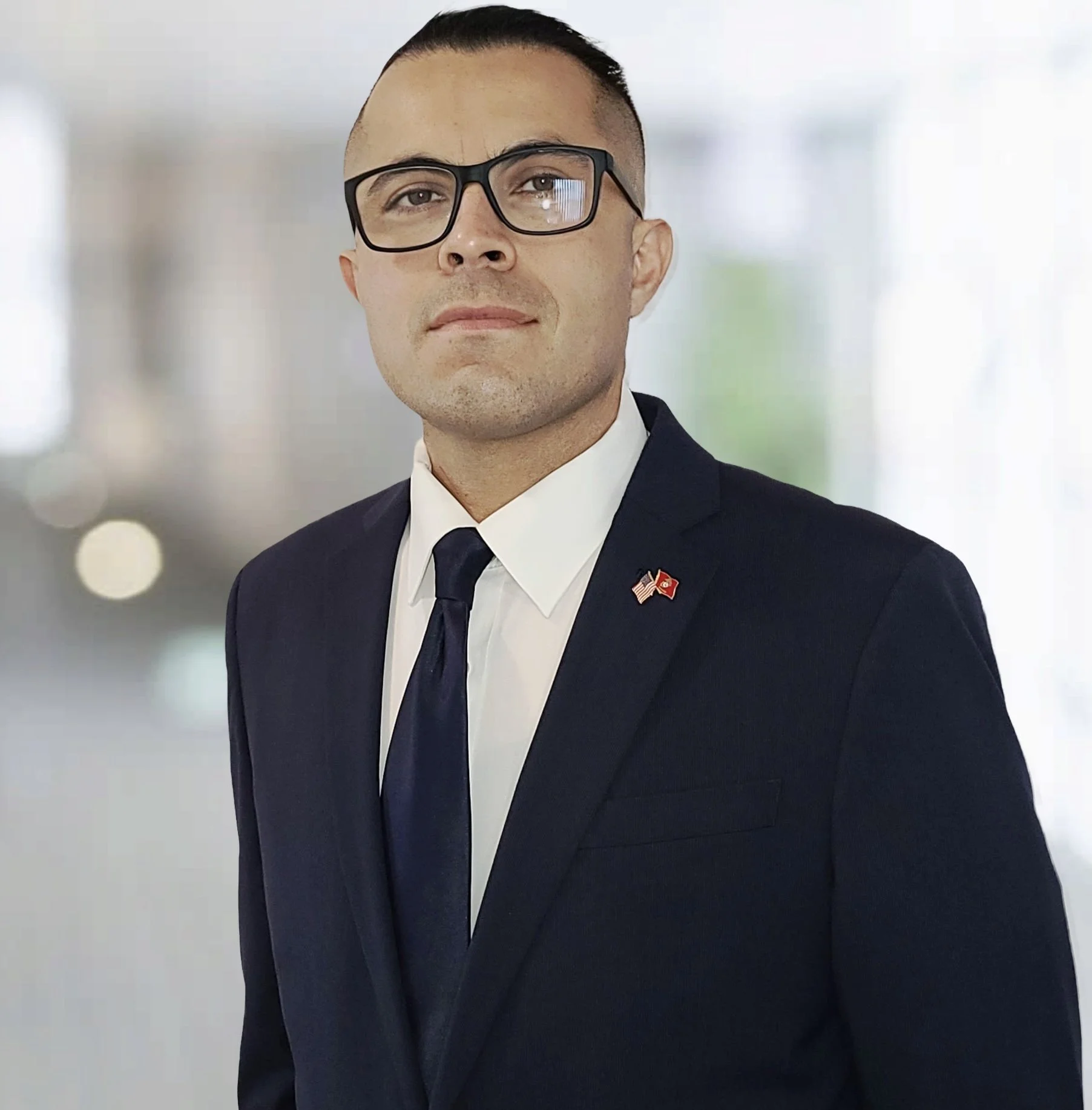 A man in a dark suit with an American and Turkish flag pin, wearing glasses, standing indoors.