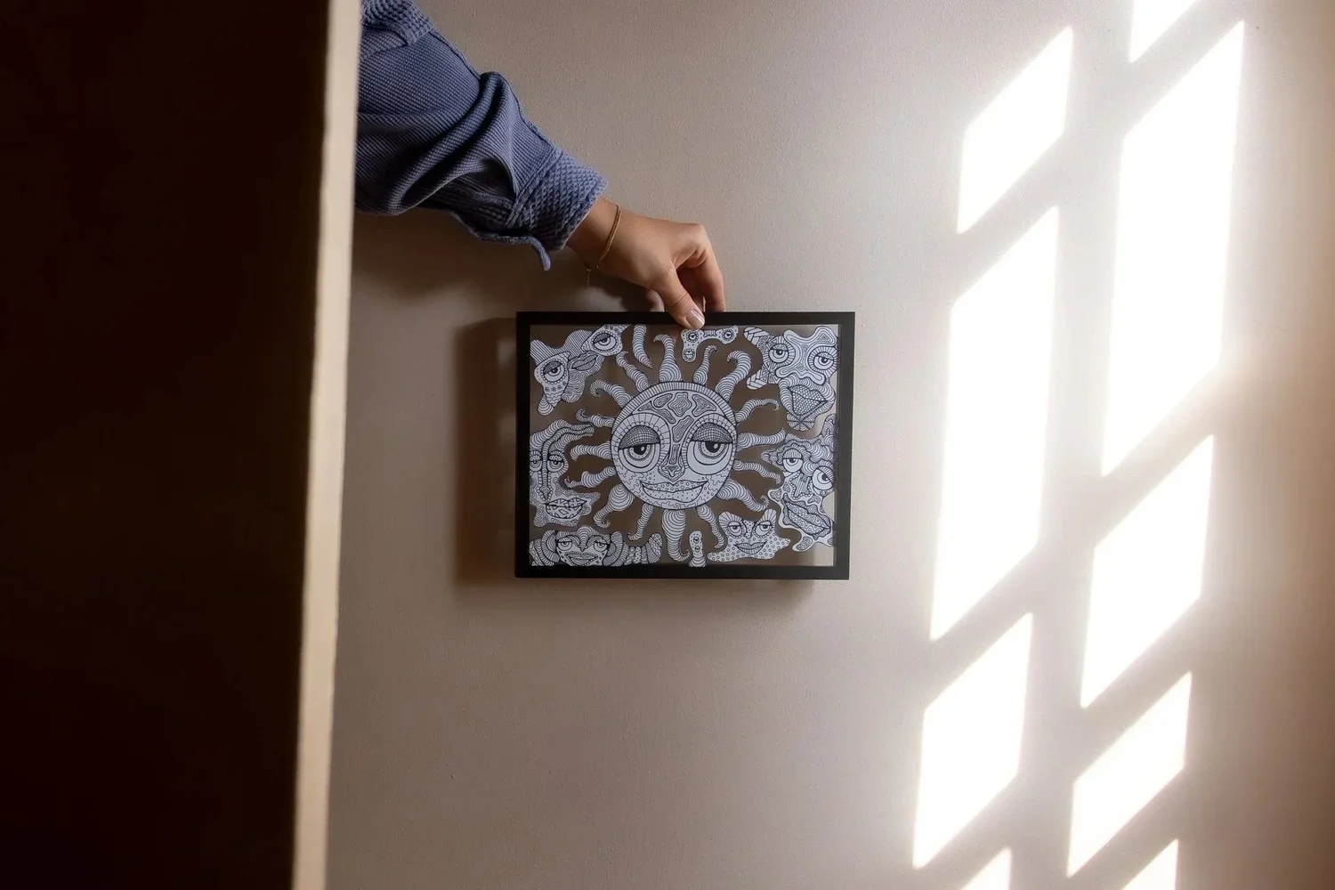 Person holding a framed black and white abstract art piece with a sun and face design, sunlight casting a patterned shadow on the wall.