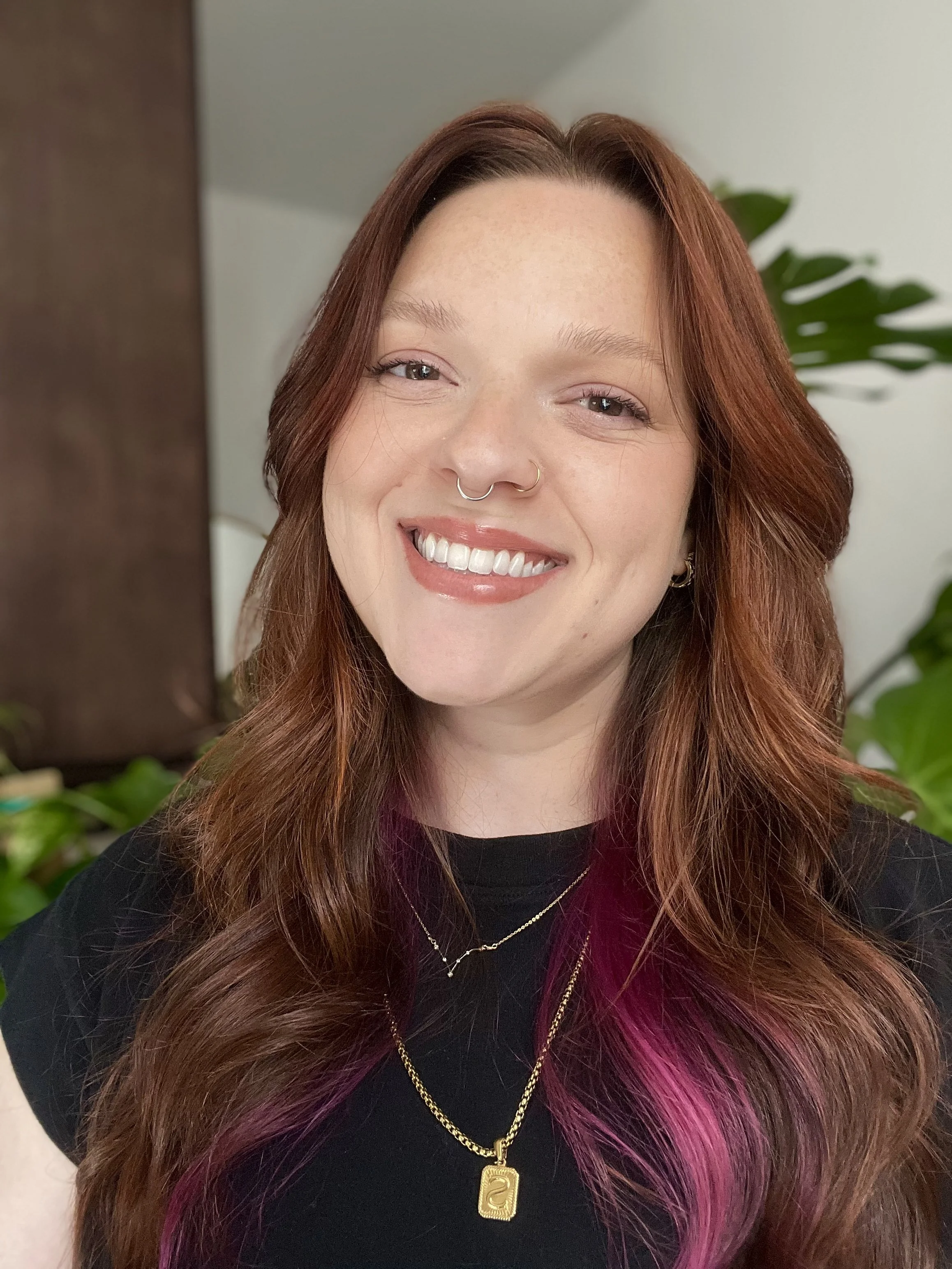 Alt text: above is an image of Stardust Therapy’s founder Stephanie who is a white, queer woman with copper and pink hair. She is posed smiling in front of a brown, and white background with plants.