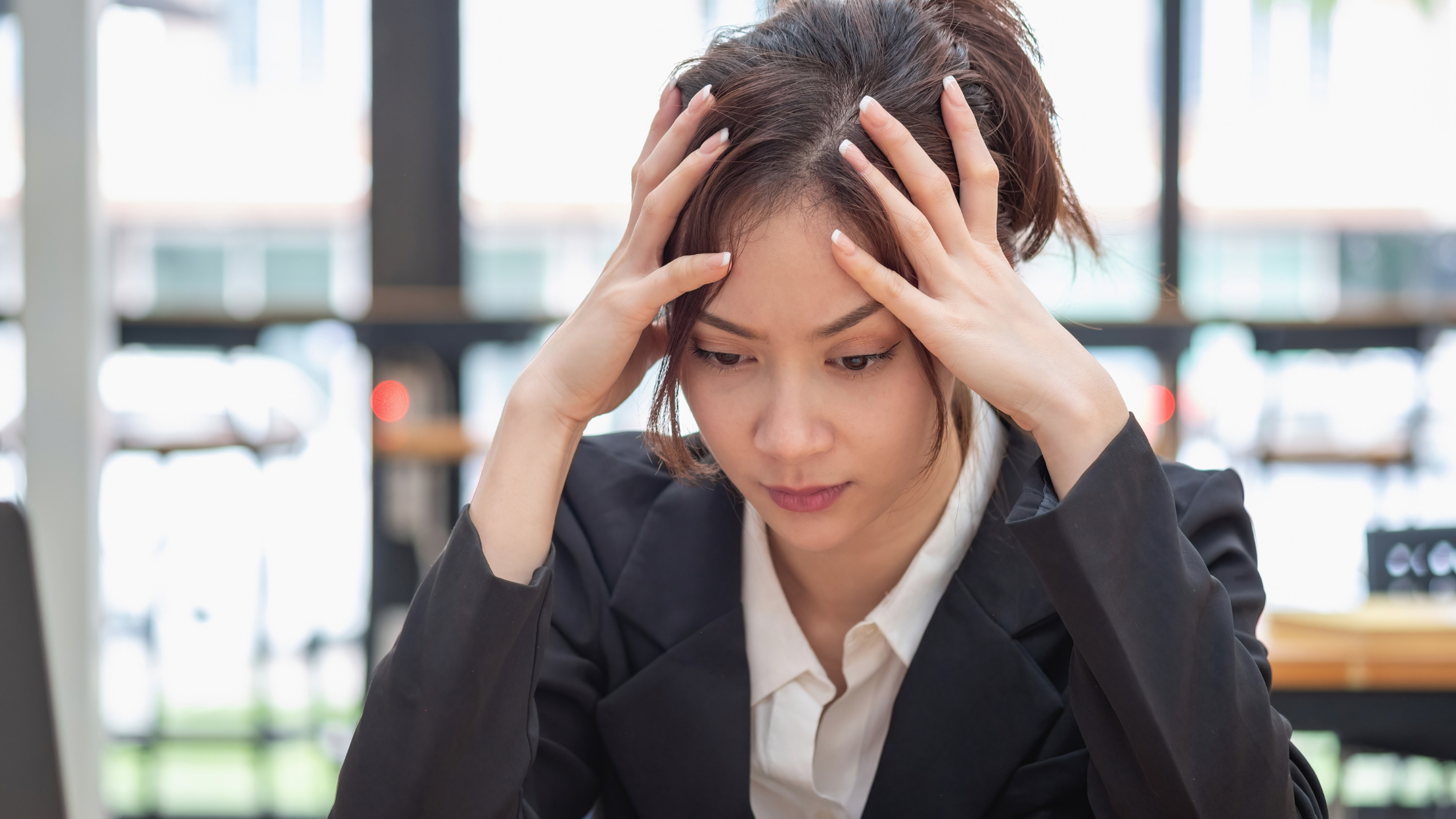Professional woman in a dark blazer holding her head in both hands at her desk, visibly overwhelmed — illustrating the emotional toll of workplace burnout
