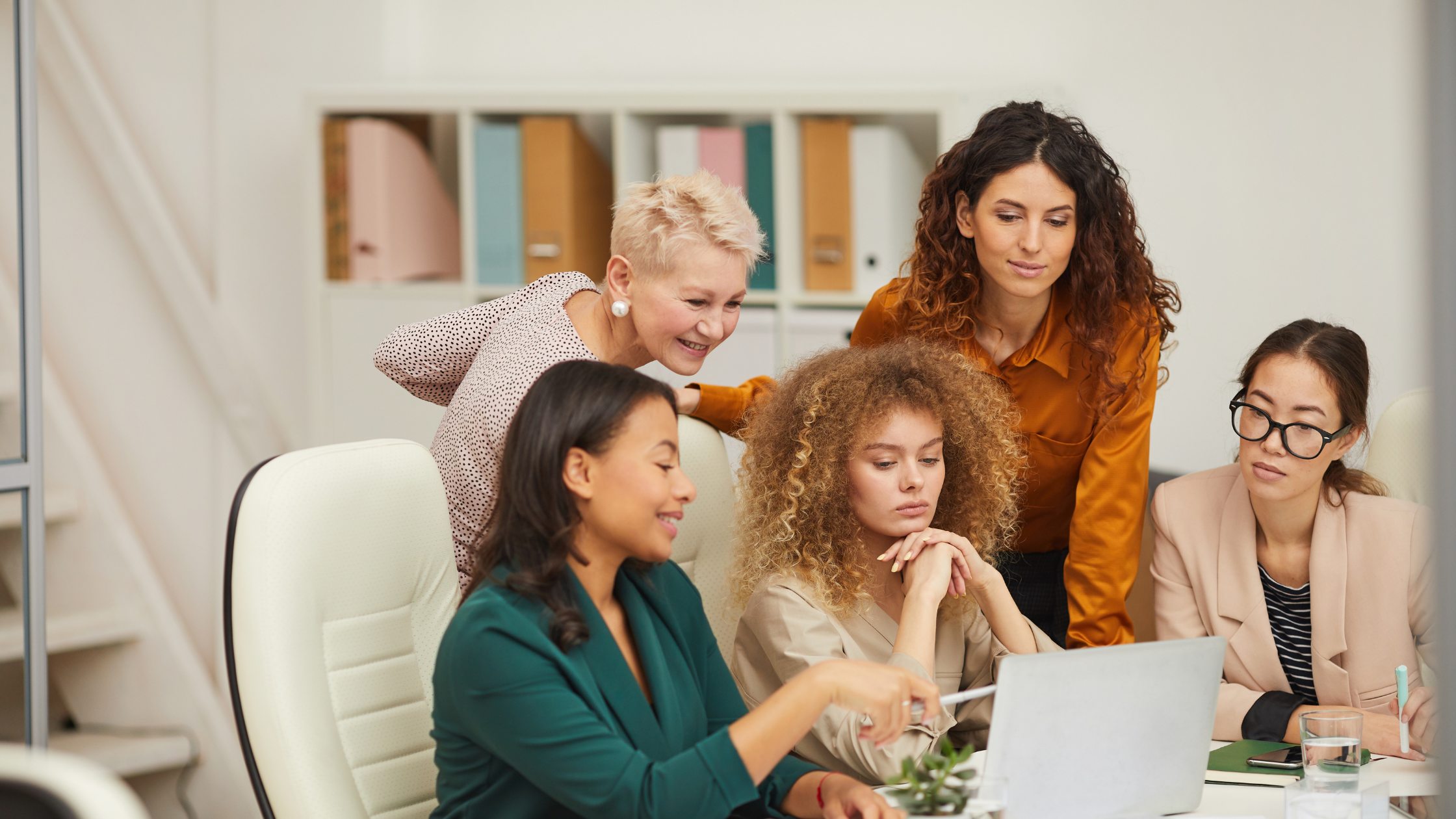 Five women collaborating around a laptop in an office, engaged in focused team discussion and shared problem-solving