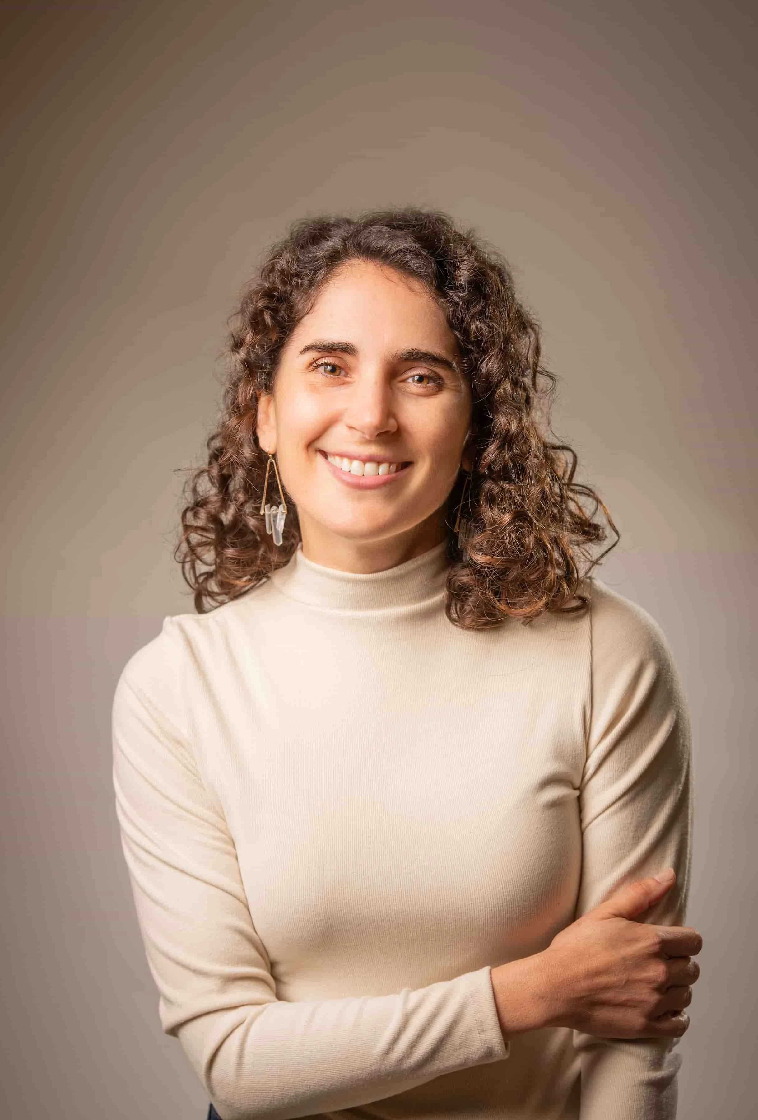 Portrait of Carly Caminiti, smiling, wearing a beige turtleneck and earrings, against a neutral background.