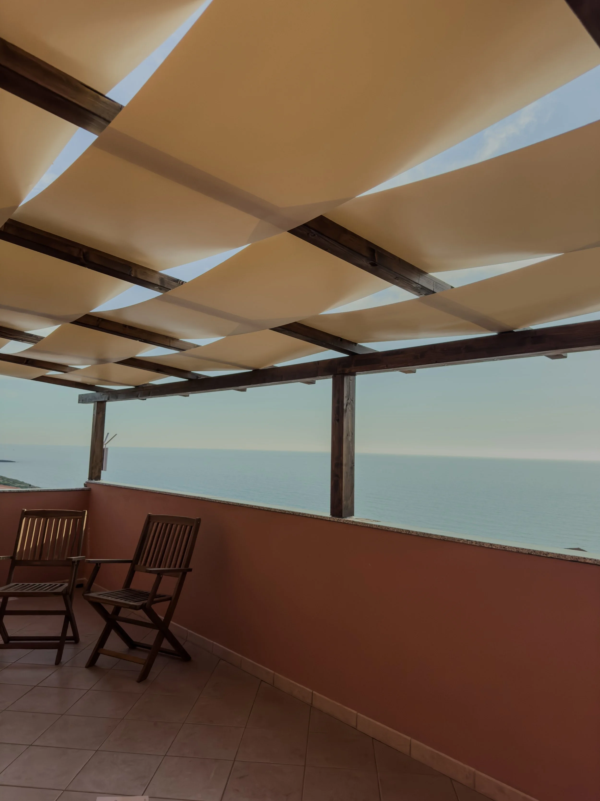 Balcony with wooden chairs and a view of the ocean, shaded by beige fabric panels on a wooden pergola.