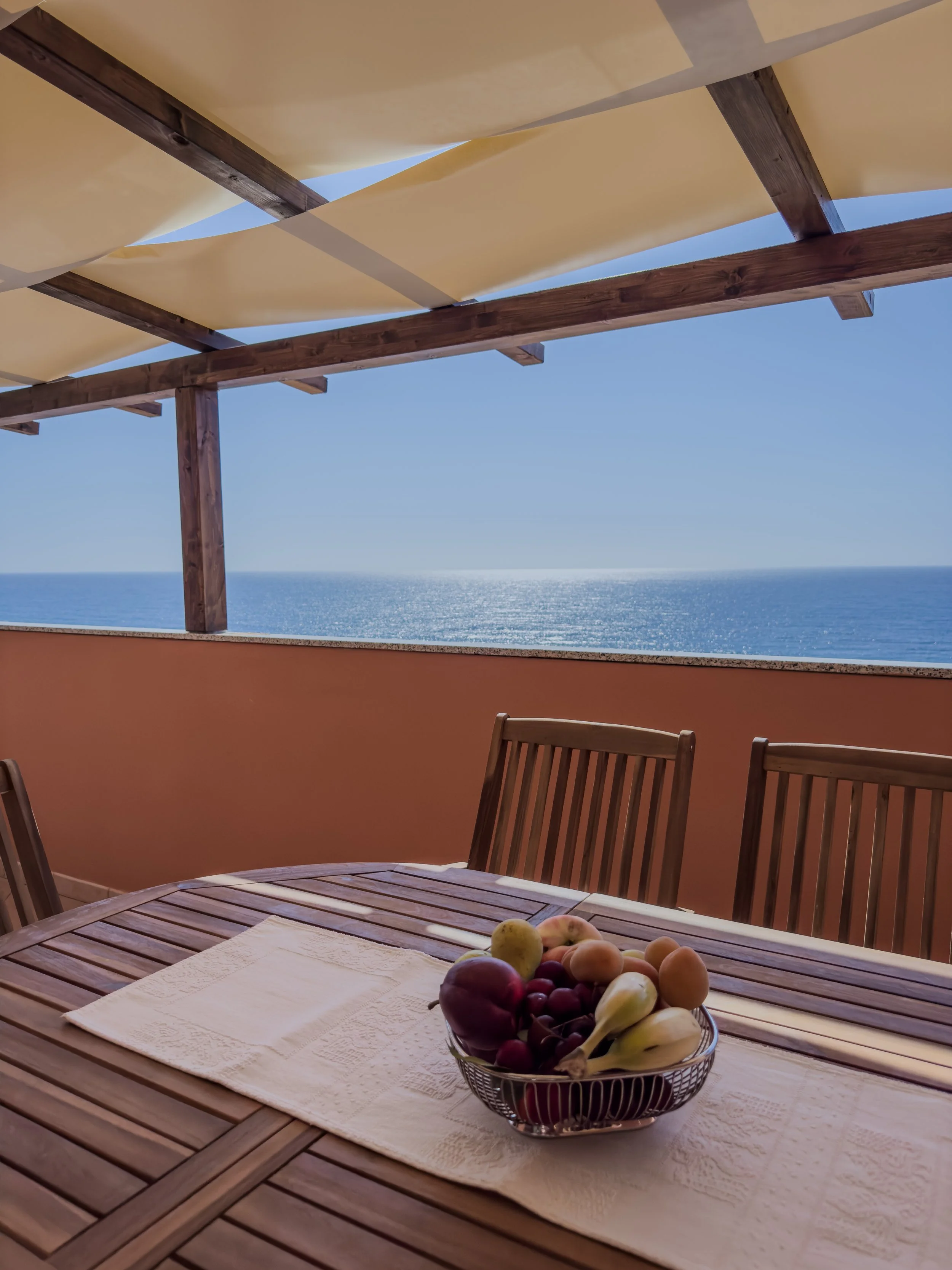 A wooden patio table with a white table runner and a bowl of assorted fruits, overlooking the ocean under a canopy with wooden beams and beige sail cloths.