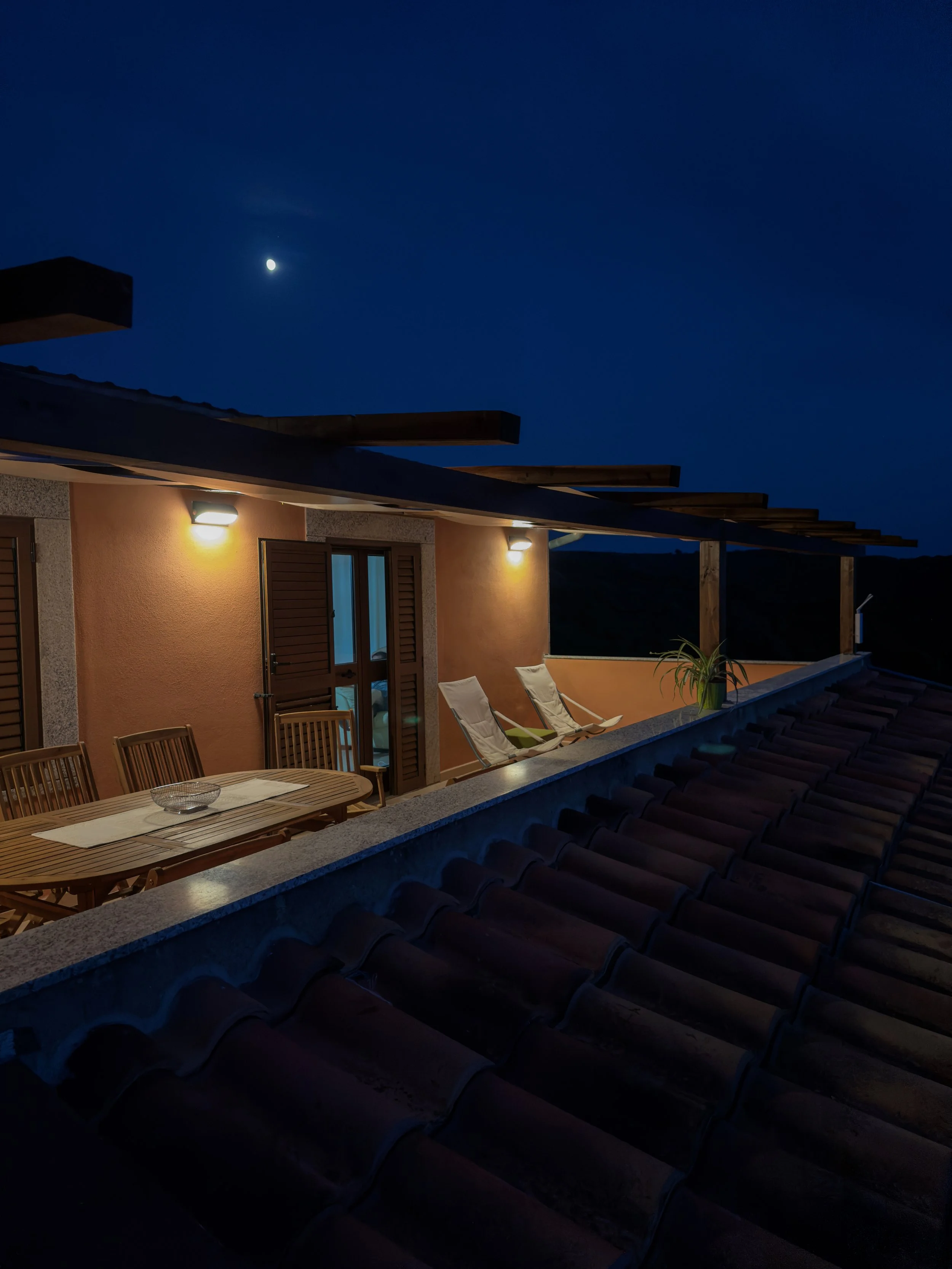 Nighttime view of a rooftop patio with a wooden dining table, chairs, lounge chairs, and potted plant, illuminated by wall lights under a dark sky with a visible moon.