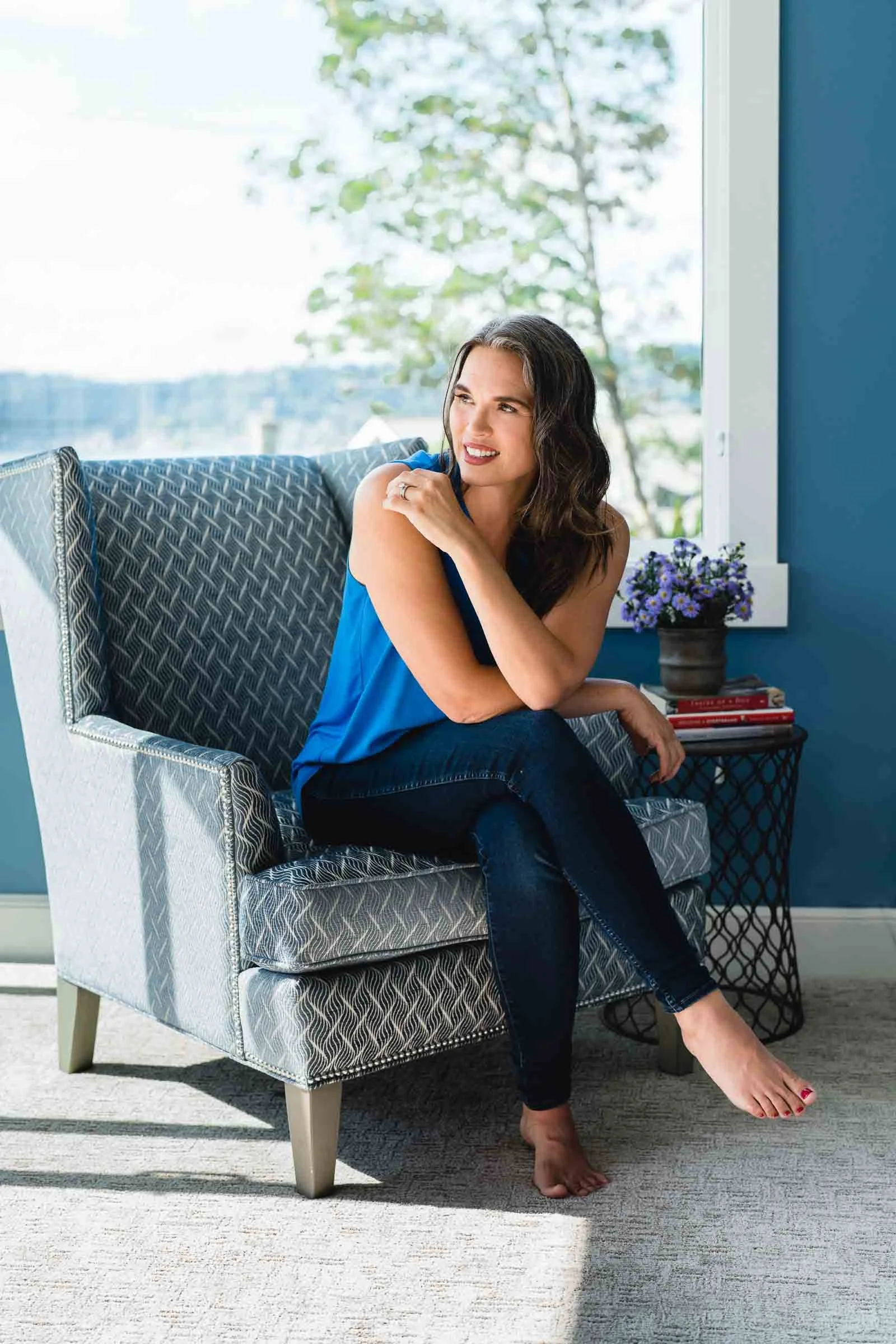 Woman sitting on a patterned armchair near a window with a view of trees, wearing a blue top and dark jeans, smiling and looking thoughtfully away.