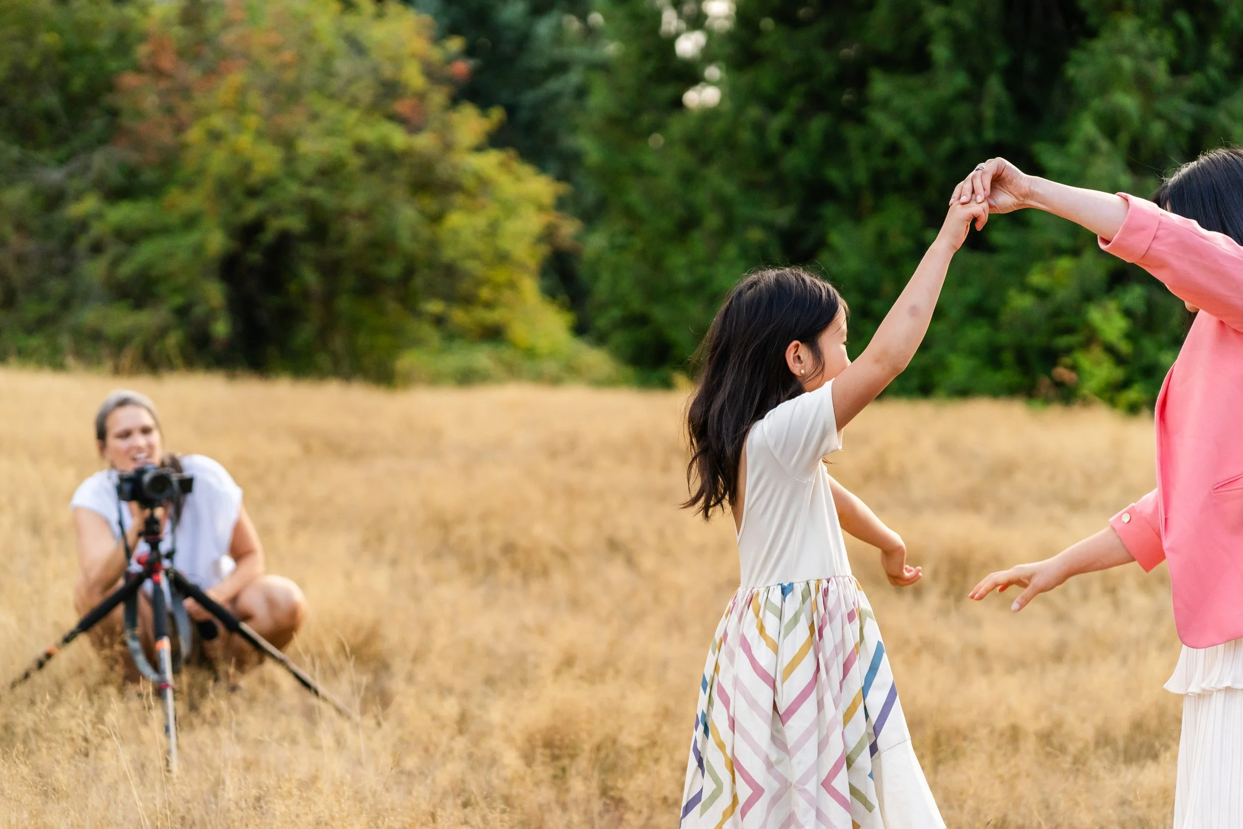 A girl dancing with two women in a grassy field with trees in the background, while a female photographer with a camera crouches nearby taking pictures
