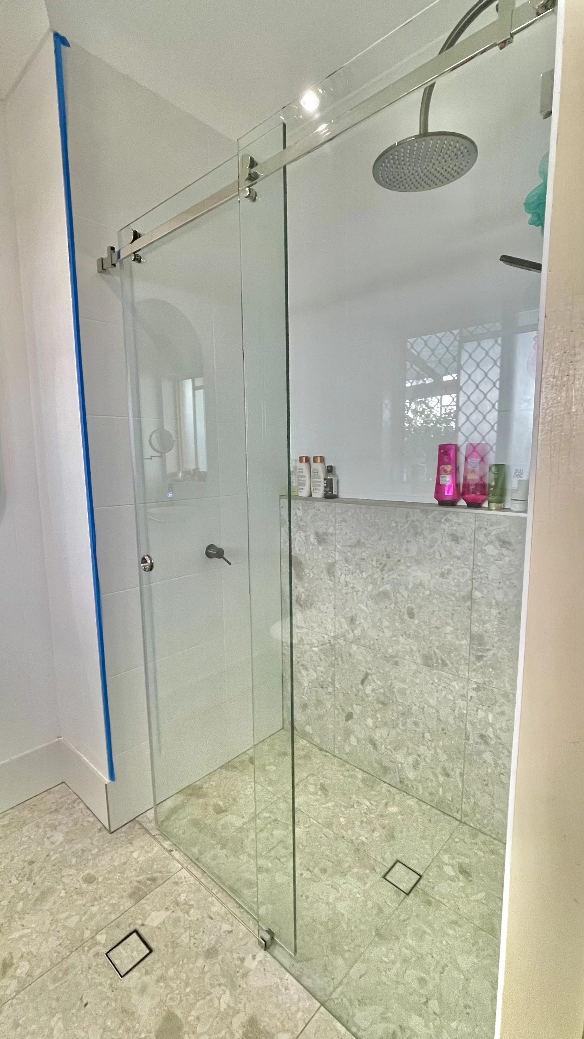 A modern shower with a glass door, a rainfall showerhead, and bottles of toiletries on a built-in shelf.