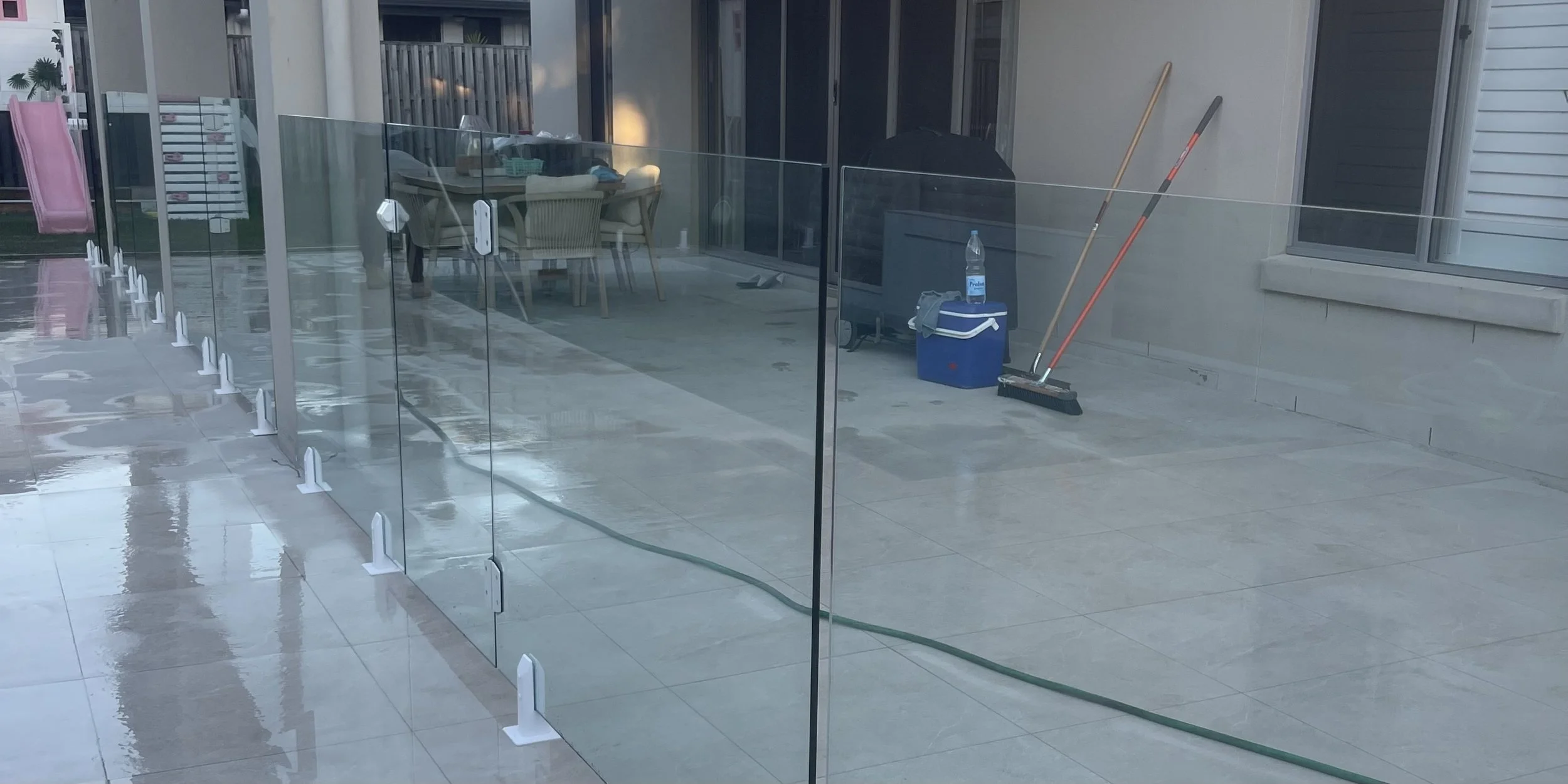 Clean glass pool fence installed on a patio with patio furniture, brooms, and a cooler visible in the background.
