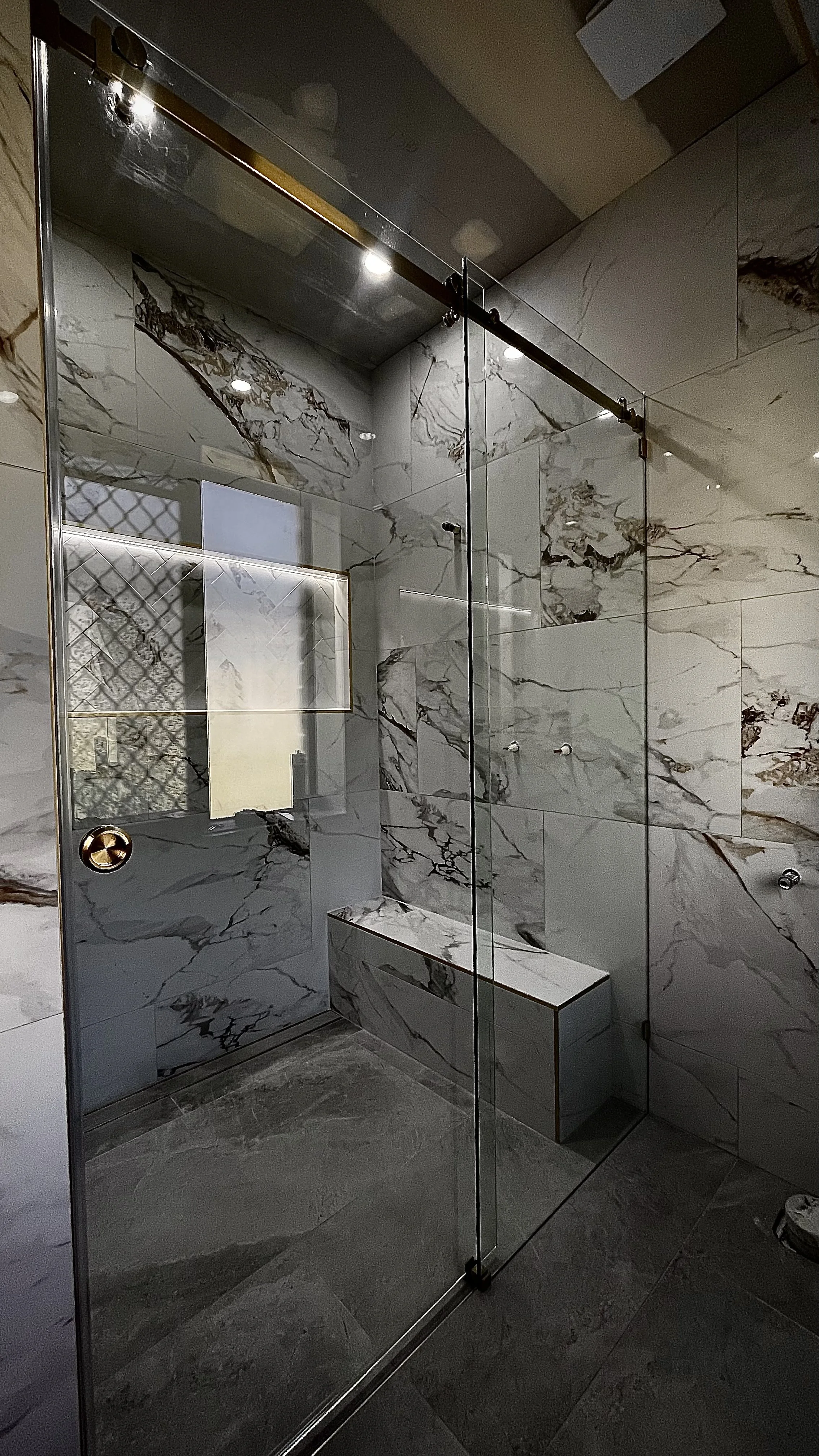 Modern shower with glass door, marble walls with brown and gray veining, gray tile floor, and built-in bench.