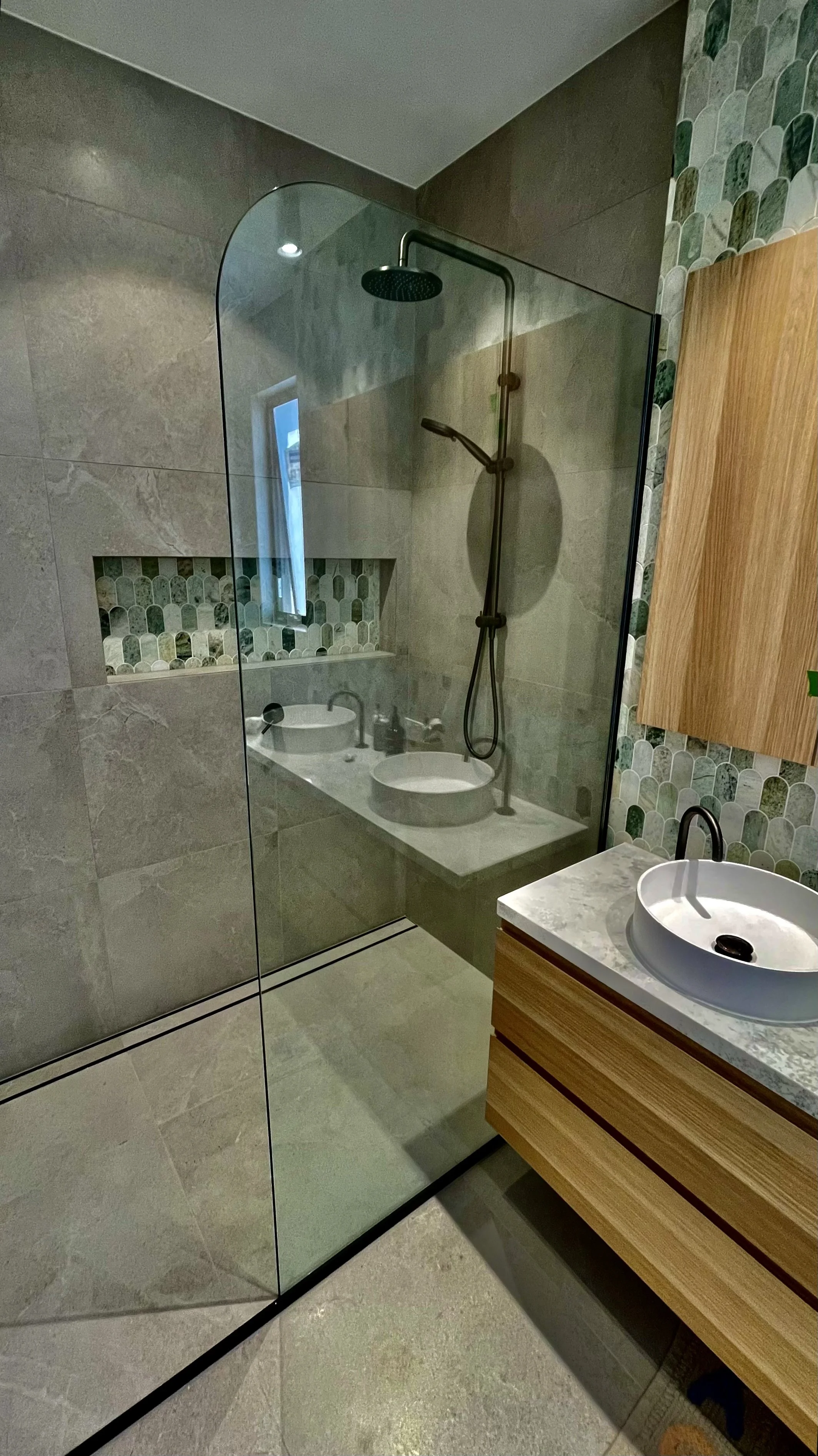 Modern bathroom shower with glass partition, rainfall showerhead, and handheld shower; bathroom vanity with vessel sink, faucet, and wooden cabinet; decorative tile wall behind sink.