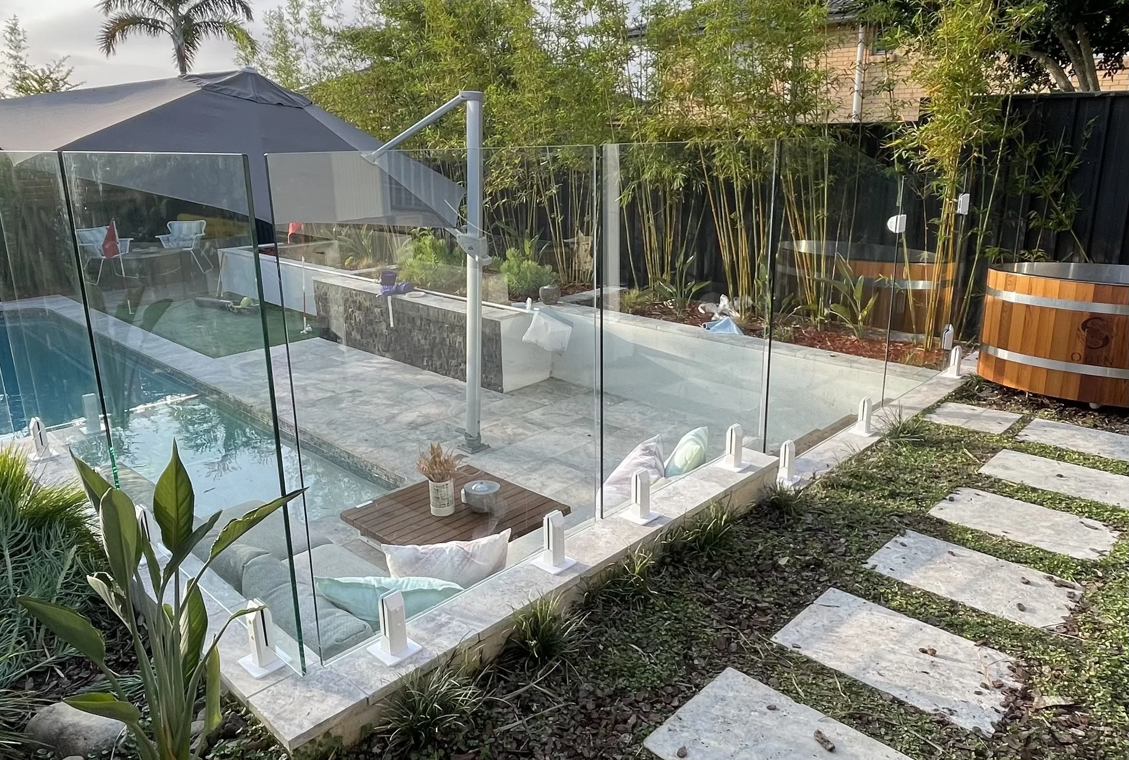 Backyard with a swimming pool enclosed by glass fencing, surrounded by greenery, with outdoor furniture and a hot tub in the background.
