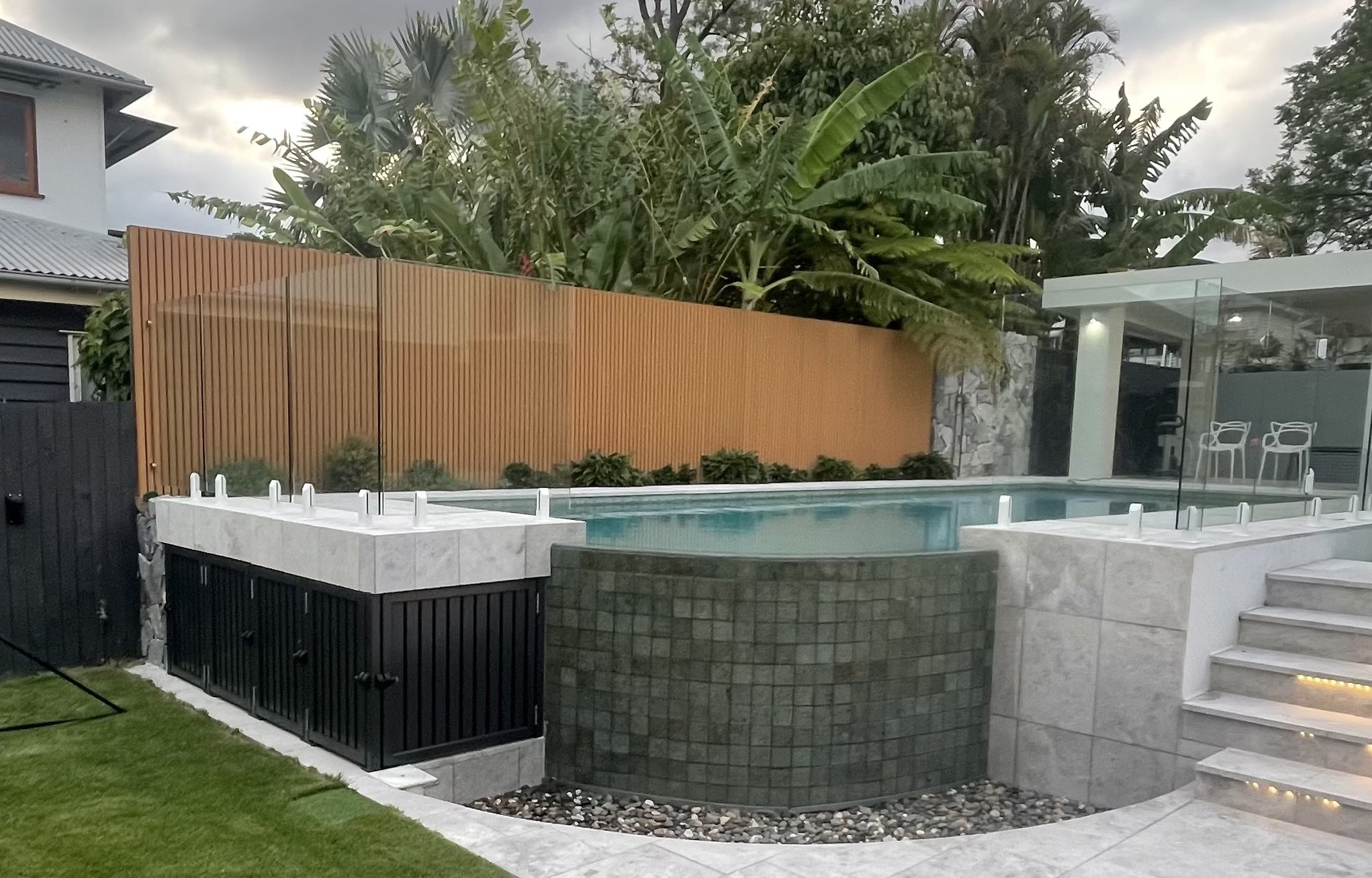 Outdoor backyard with a pool, a modern glass fence, stairs with built-in lighting, a wooden privacy fence, lush green plants, and a seating area with chairs, during cloudy weather.