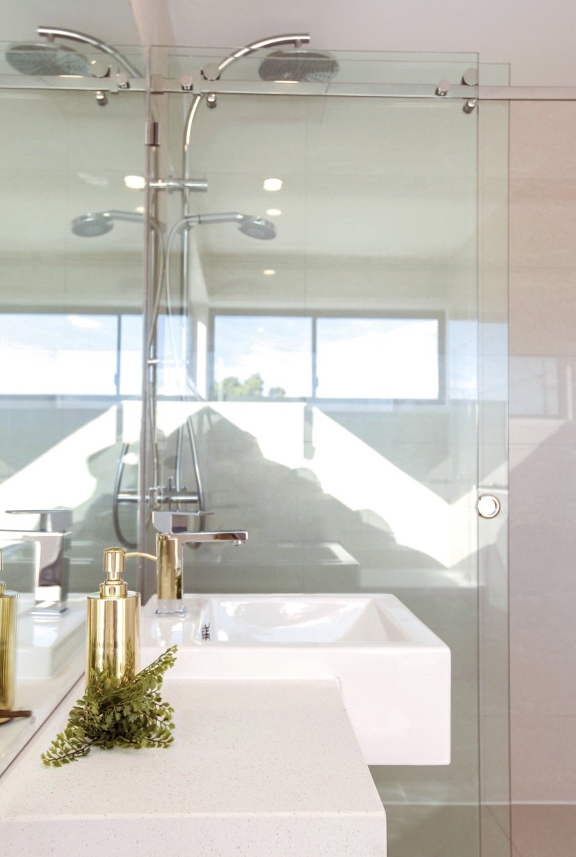 Modern bathroom with a glass-enclosed shower, dual shower heads, a white vanity with a sink, and gold soap dispensers, with sunlight coming through large windows.