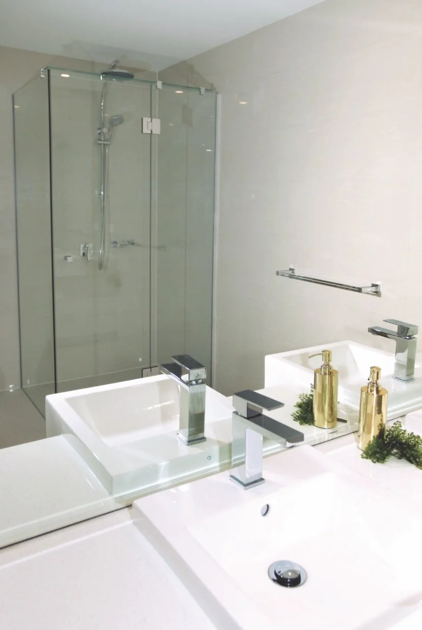 Bathroom with two white sinks, mirrors, gold soap dispensers, and a glass shower enclosure in the background.