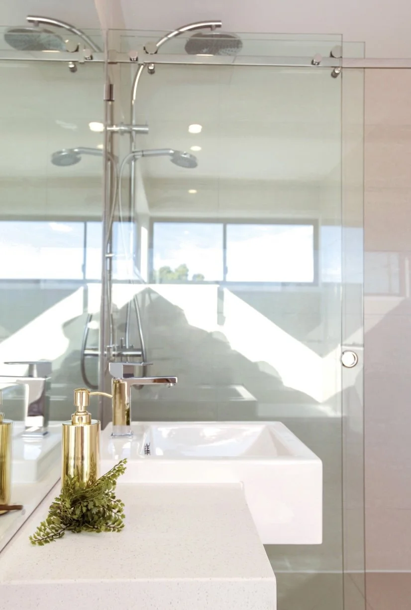 Modern bathroom with a white countertop, gold soap dispenser, and a small green plant, reflected in a large mirror behind it. A glass shower enclosure with silver fixtures and a showerhead is visible in the background.