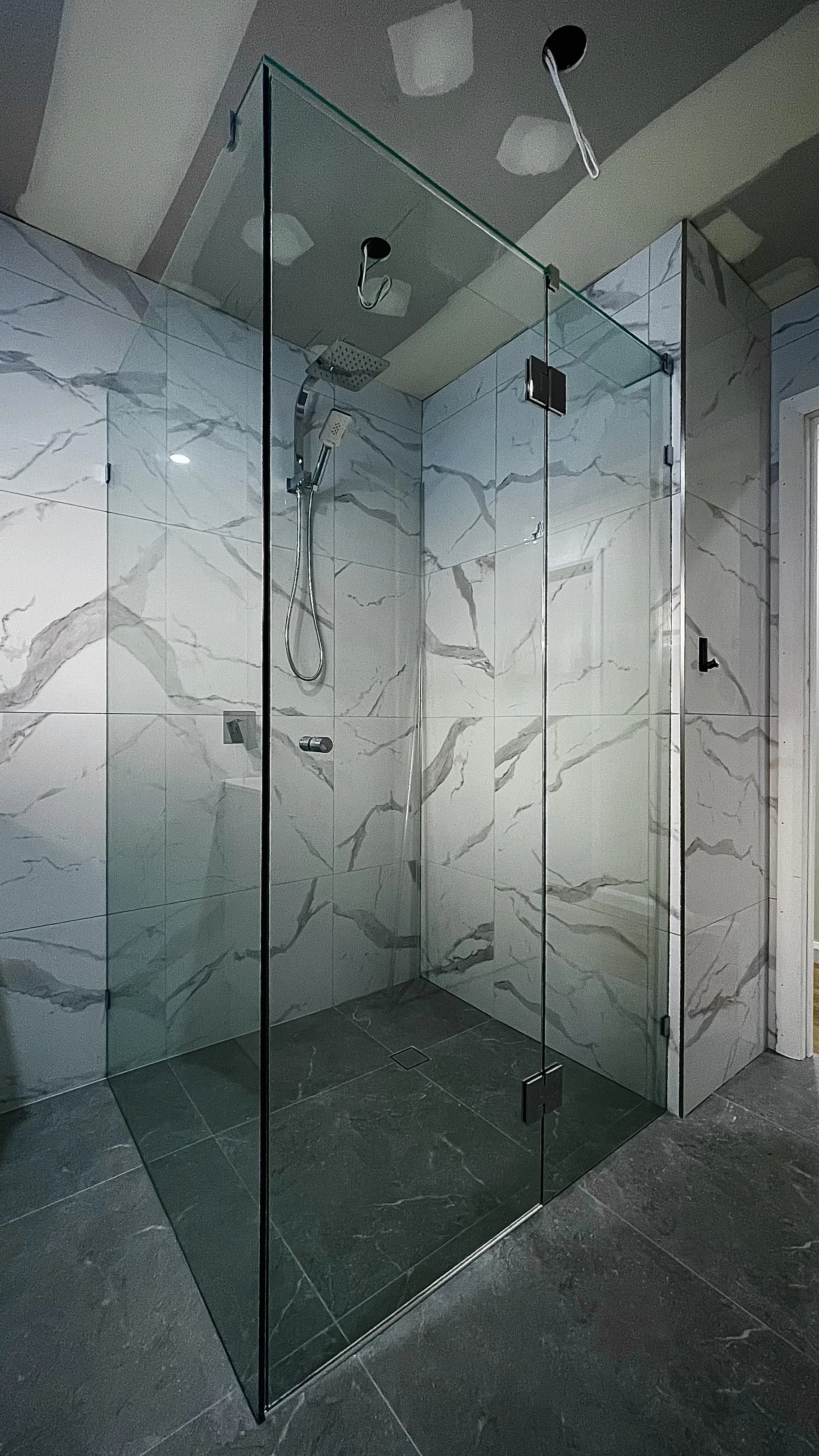 Modern glass shower enclosure with marble wall tiles and dark gray floor tiles, containing a rainfall showerhead and handheld shower.