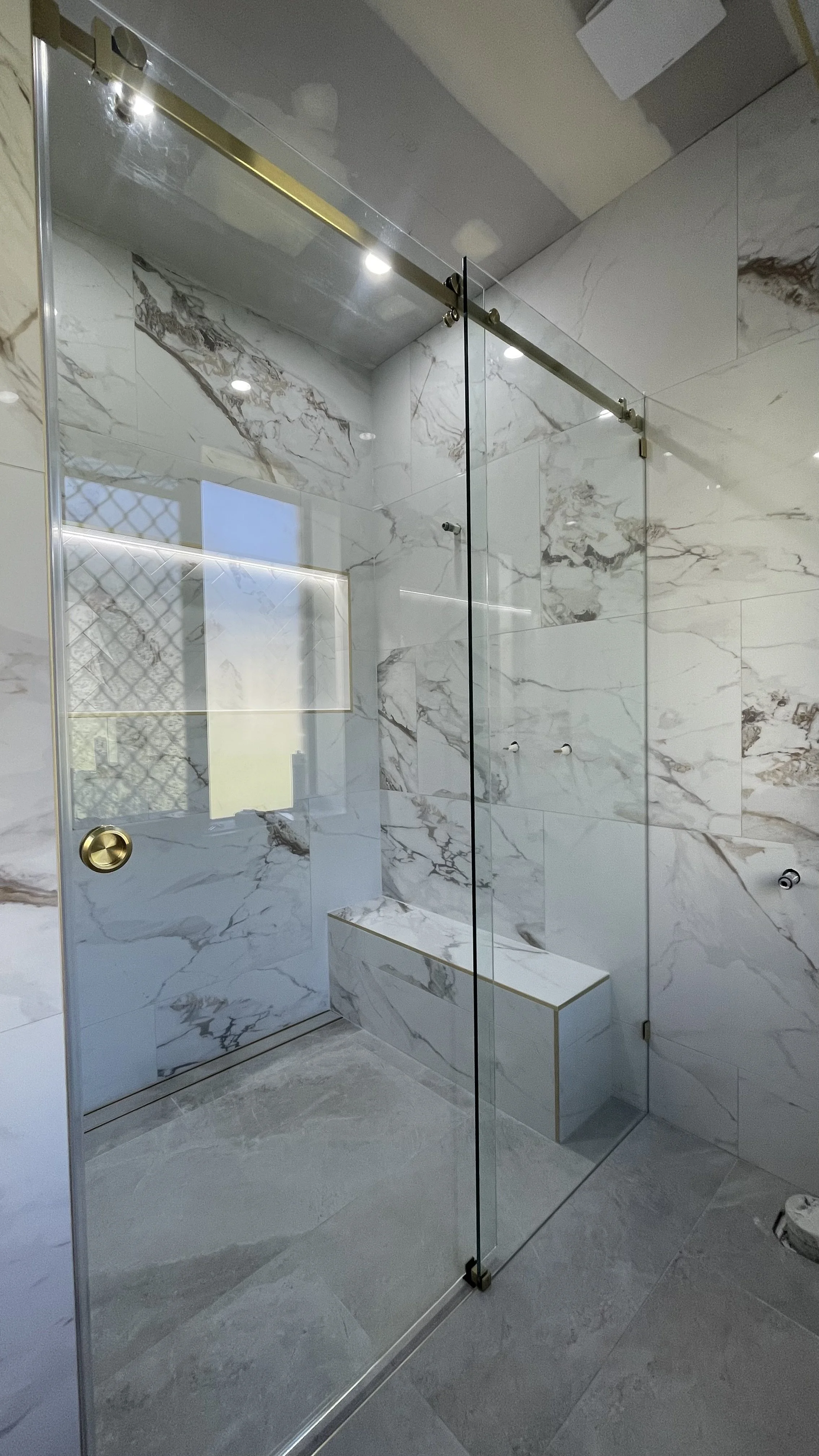 Modern walk-in shower with marble walls and bench, glass enclosure, gold fixtures, and grey tile floor.