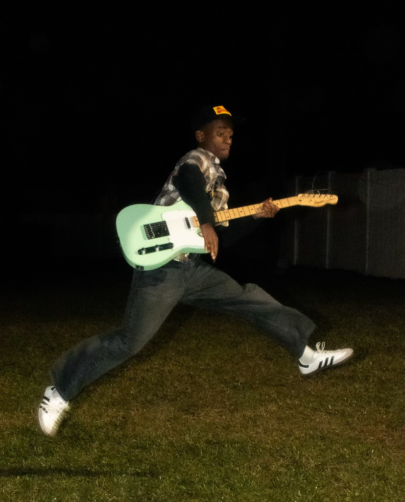 Person in casual clothes and a cap jumping on grass at night while holding a mint green electric guitar.