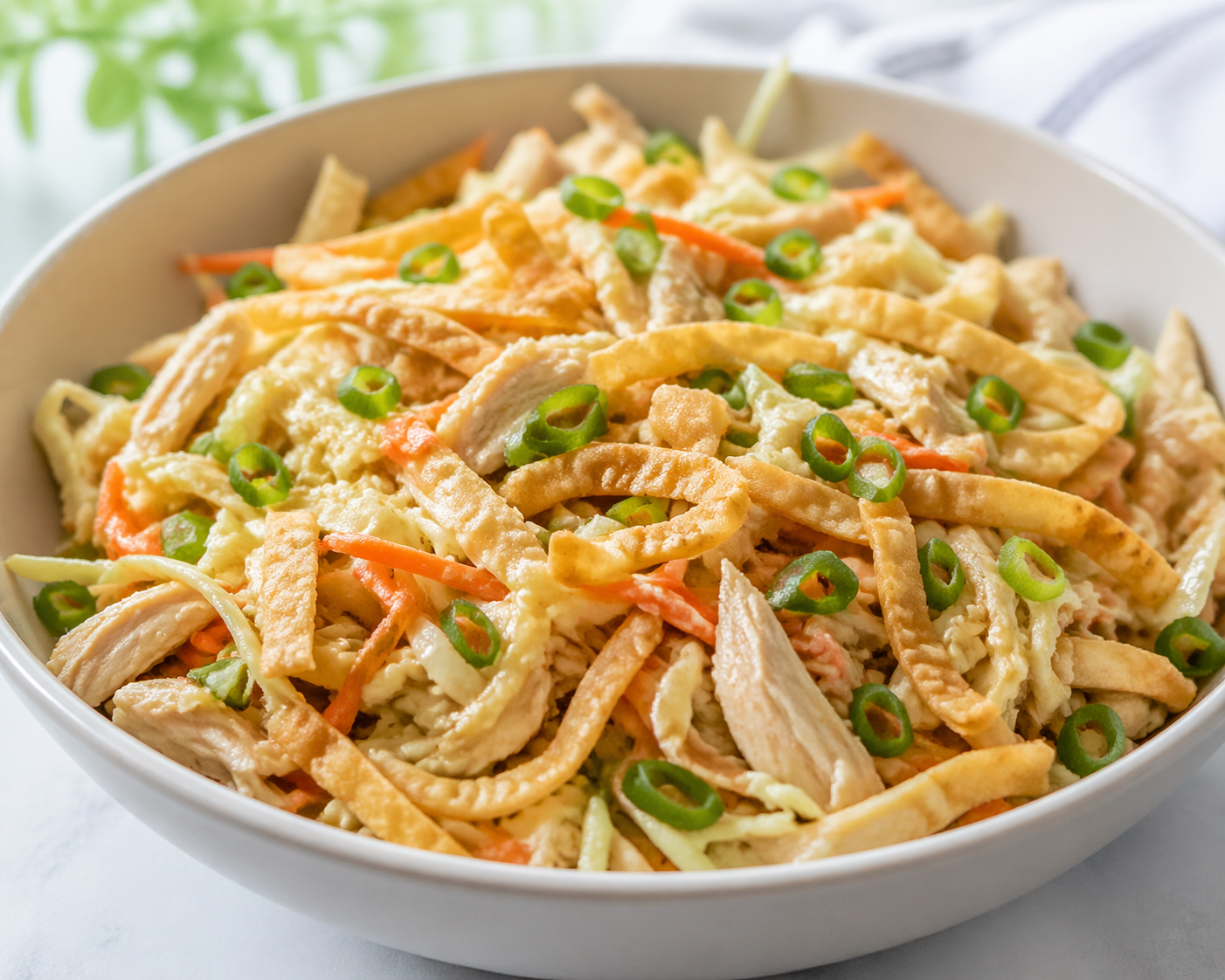 Chicken wonton crunch salad with cabbage, shredded carrots, sliced green onions, crispy wonton strips, and sesame dressing in a white bowl