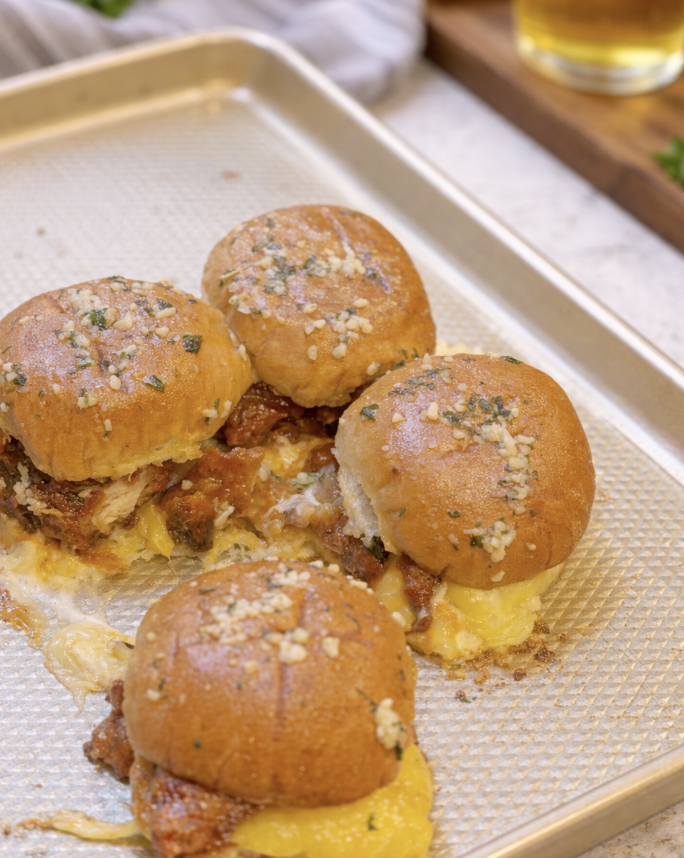 Hot honey chicken sliders on garlic butter Hawaiian rolls baked on a sheet pan with melted cheese and savory sauce.