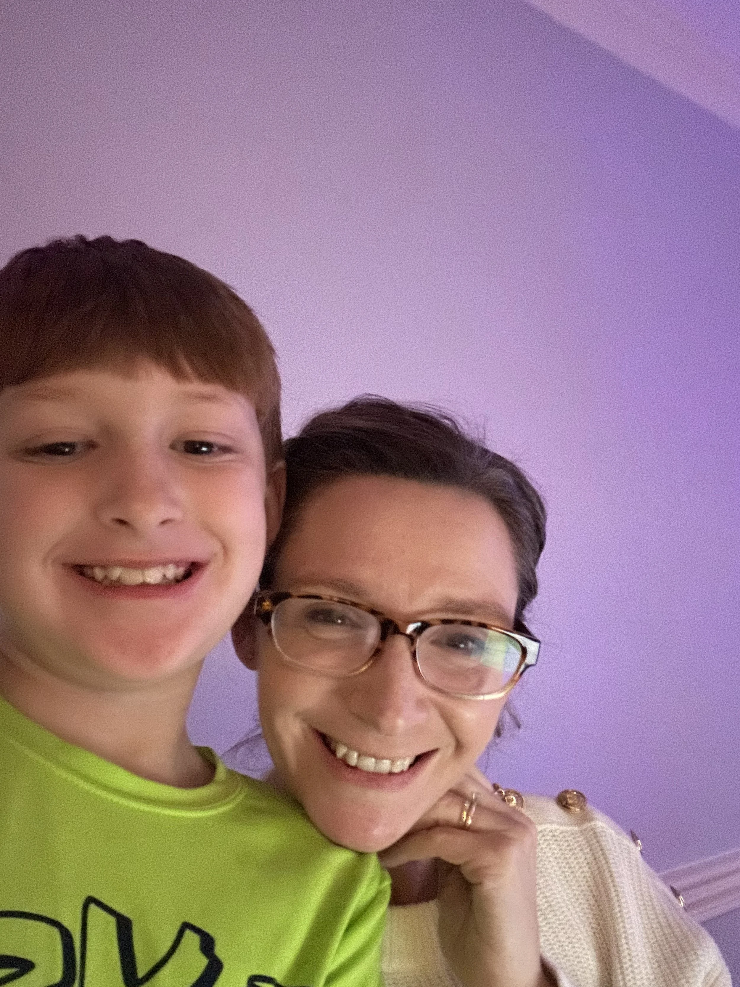 A smiling woman and a young boy taking a selfie together, with a purple ceiling in the background.