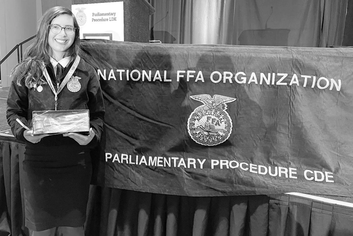 A young woman in a FFA jacket holding a plaque, standing next to a banner that reads 'National FFA Organization' and 'Parliamentary Procedure CDE' with the FFA emblem.