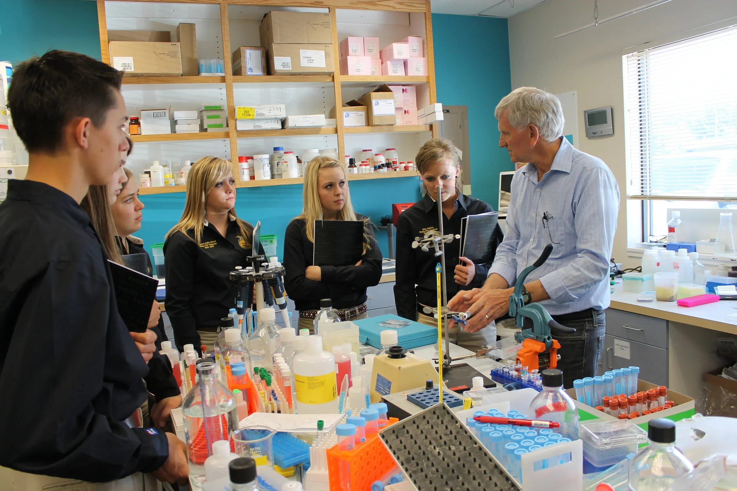 A classroom or laboratory setting with a group of students gathered around a lab table, listening to an older male instructor who is explaining scientific or laboratory equipment. The table is filled with various laboratory supplies such as test tubes, bottles, and pipettes.