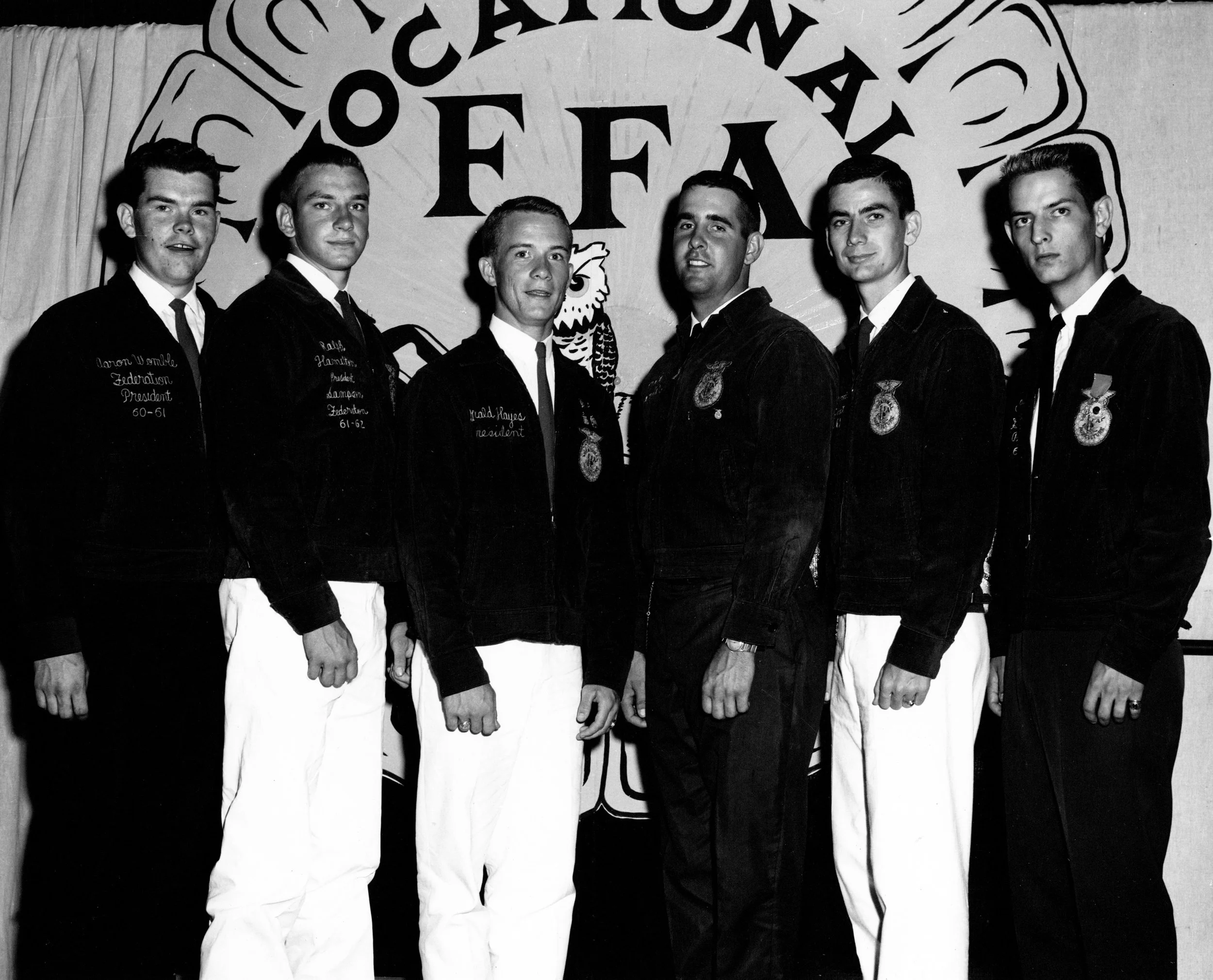Black and white photo of six young men in FFA jackets standing in front of an FFA banner, posing for a group photo at an FFA event.