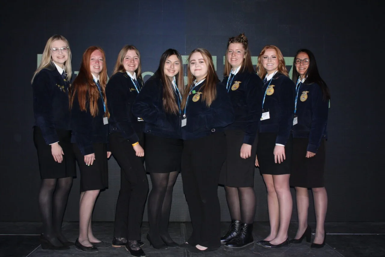 Group of eight young women wearing navy blue jackets with FFA emblems, standing together on a stage.