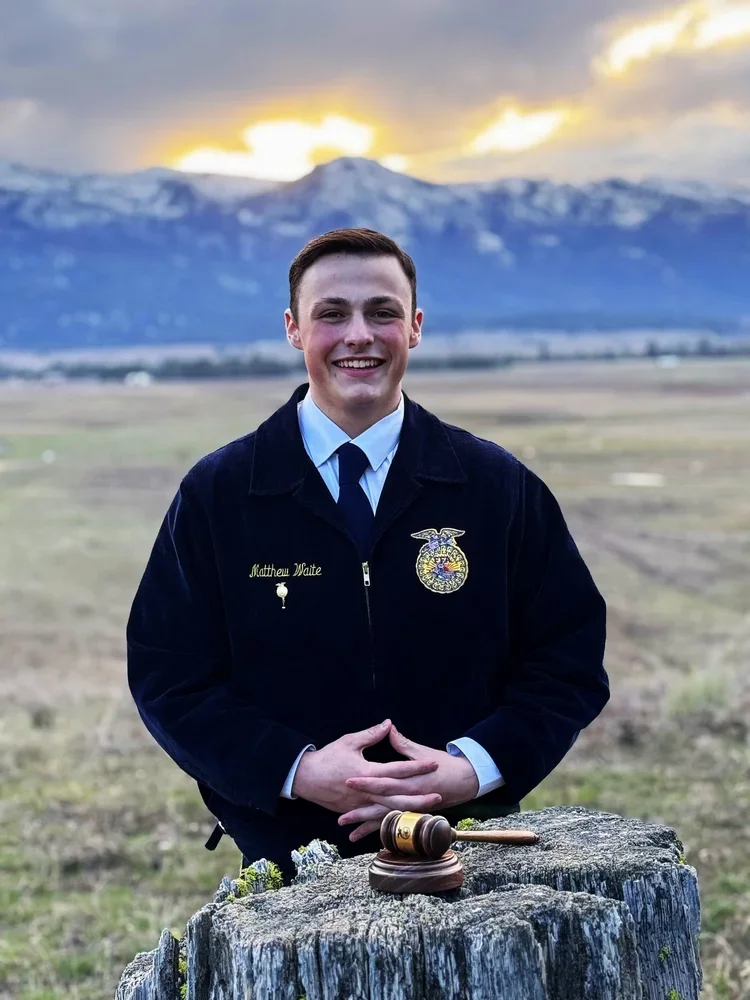 Young man dressed in a FFA uniform standing outdoors near a tree stump with a gavel on top, smiling with mountains and a cloudy sky in the background.