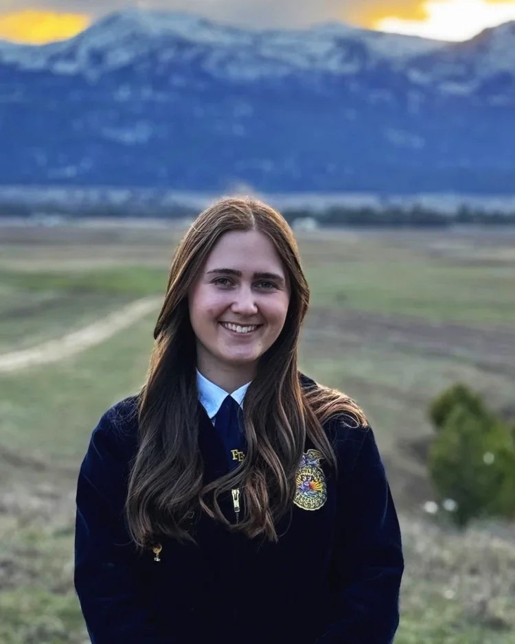 A young woman with long brown hair smiling outdoors, wearing a navy blue jacket with a zipper and patch, with a scenic mountain landscape and cloudy sky in the background.