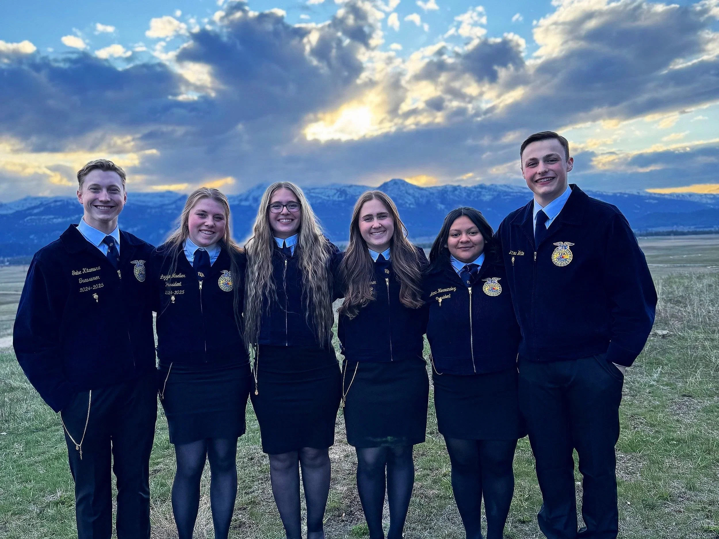 Group of six young people in FFA jackets standing outdoors in a grassy field with mountains and a cloudy sky in the background.