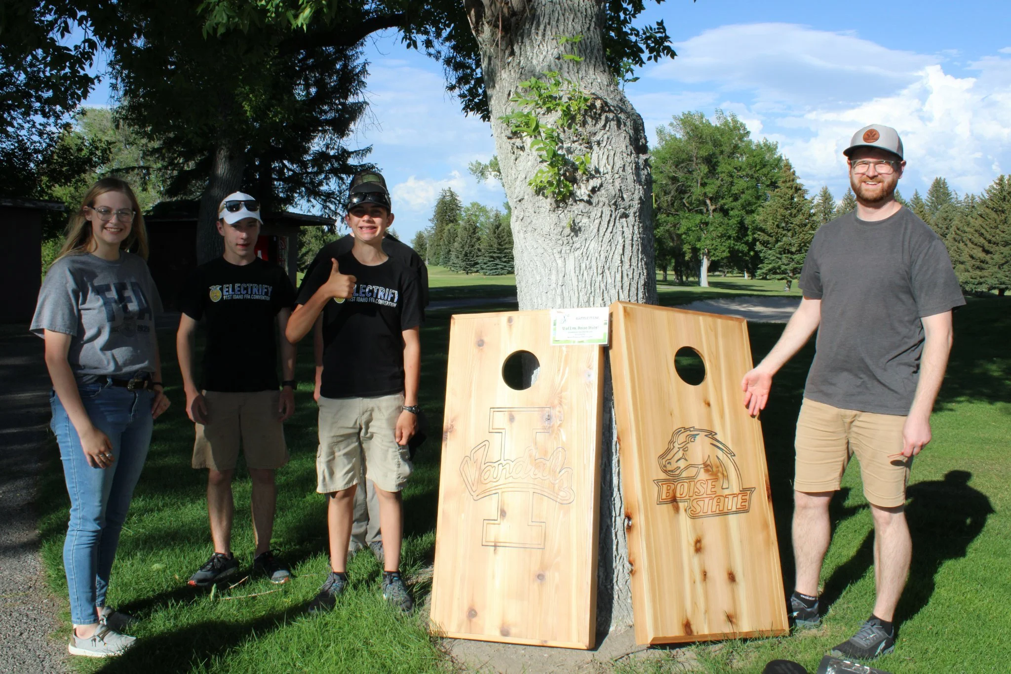 Four people standing outdoors near a tree with two wooden cornhole game boards with University of Idaho and Boise State logos leaning against the tree.