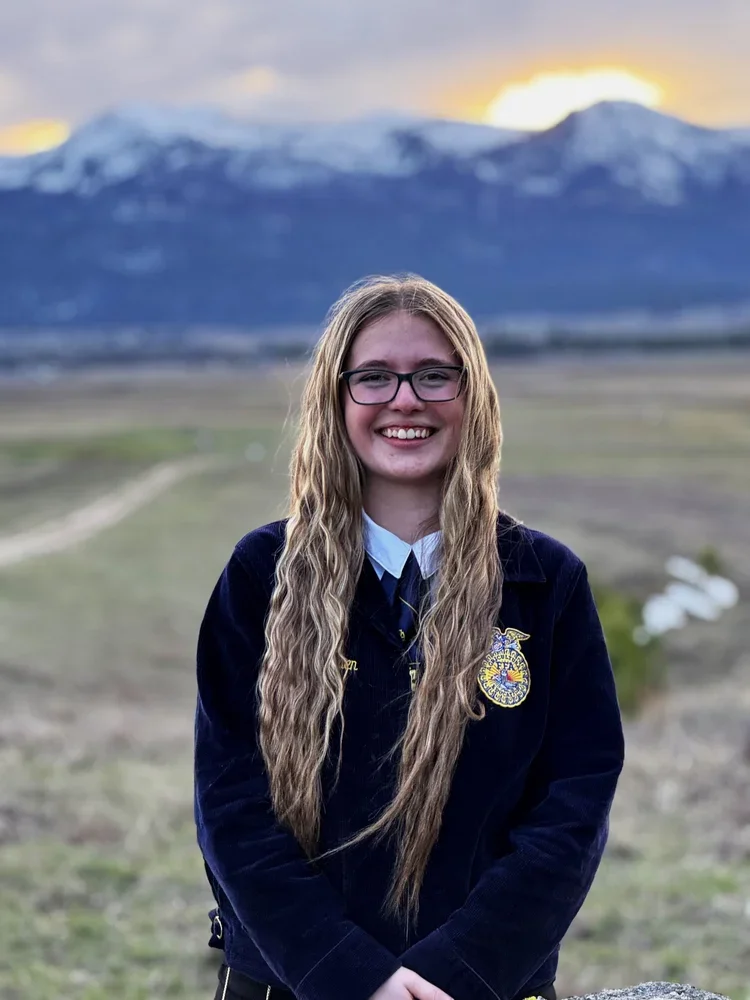 A young woman with long wavy blonde hair and glasses smiling outdoors in a field with mountains and snow at sunset in the background.