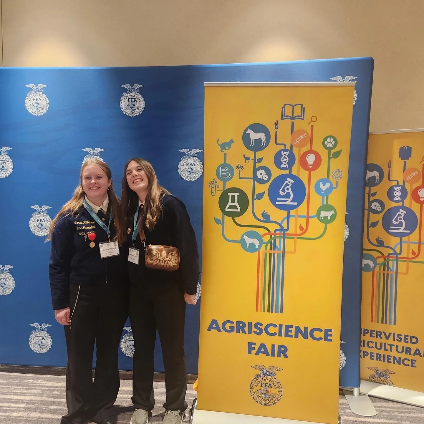 Two young women smiling and posing for a photo at the Agriscience Fair, standing next to a yellow banner with graphics of various science and agriculture symbols and the event name.