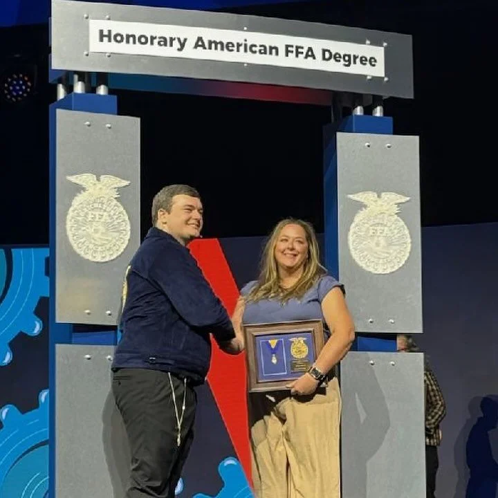 Two people smiling and shaking hands on stage during an award ceremony. The woman is holding a framed statement of achievement, and the man appears to be congratulating her. The stage background includes large emblem symbols and a banner that reads 'Honorary American FFA Degree.'