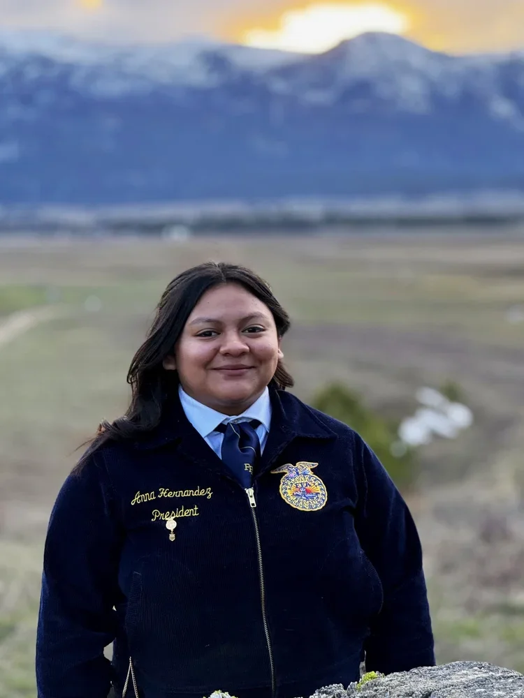A woman in a navy jacket with a FFA emblem and the name Anna Hernandez, standing outdoors with mountains and a cloudy sky in the background.