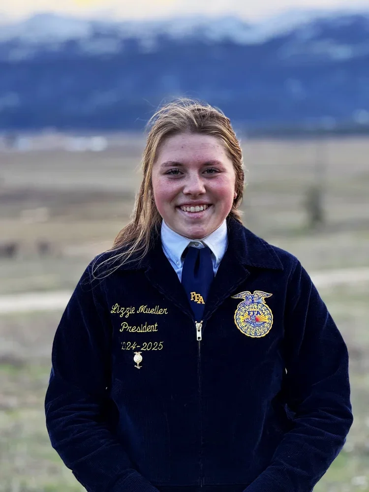 Young woman smiling outdoors, wearing a navy FFA jacket with embroidery and a tie, with mountains and open land in the background.