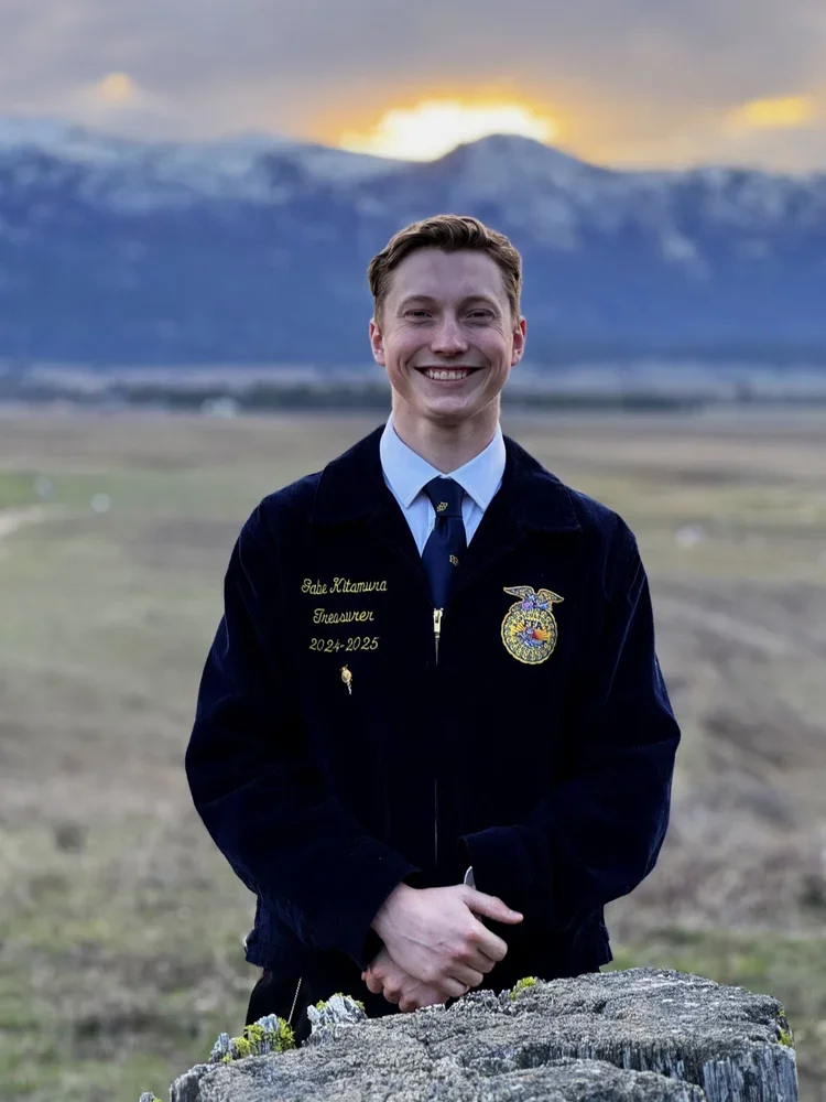 Young man in a FFA jacket standing outdoors with mountains and a sunset in the background.