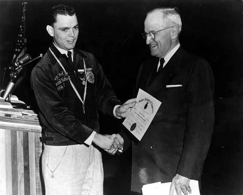 A young man in a jacket receiving a certificate from an older man in a suit during a formal ceremony, with an American flag in the background.
