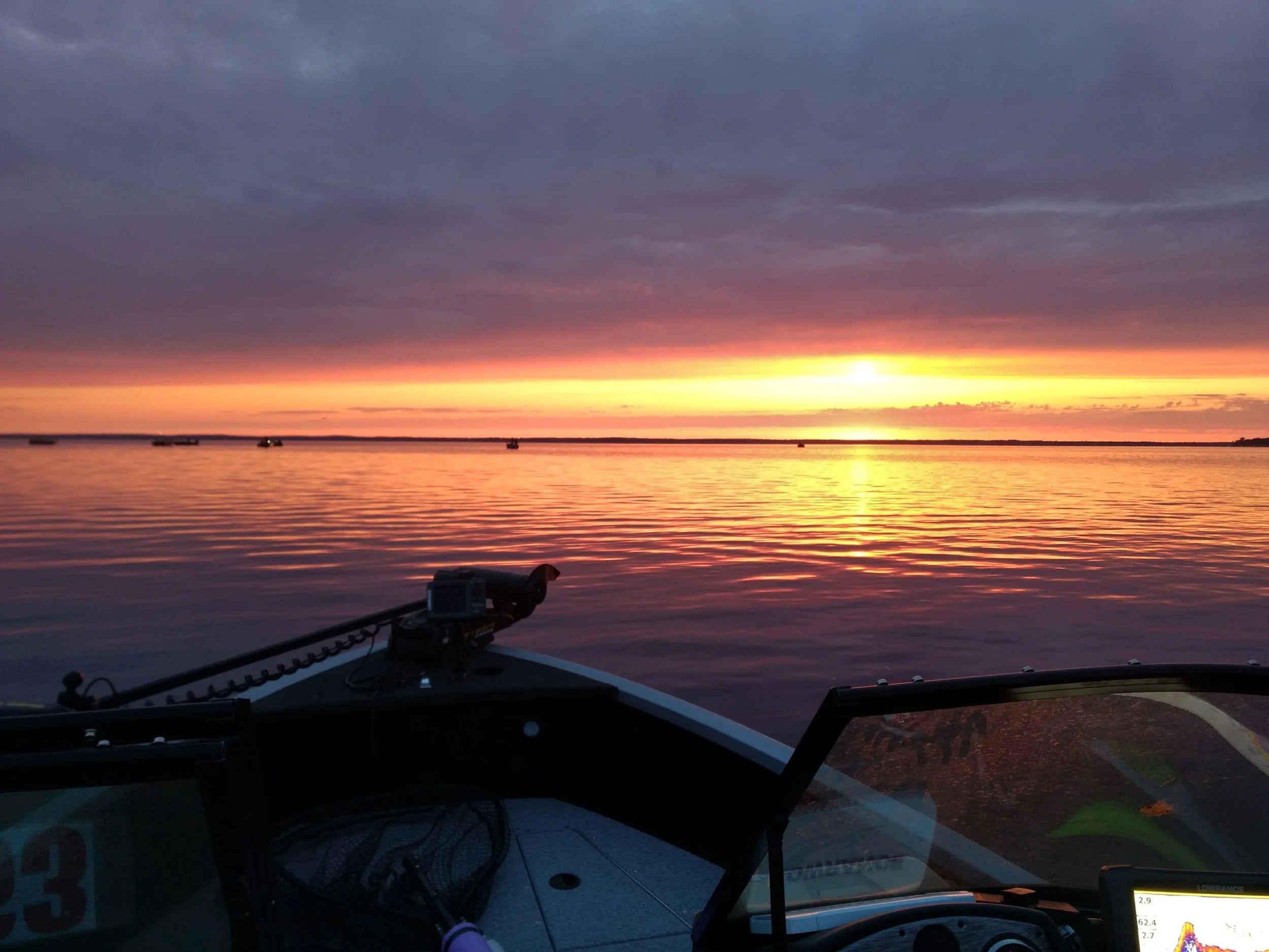 Sunset over a calm lake viewed from a boat, with boats in the distance and colorful sky with orange, purple, and pink hues.