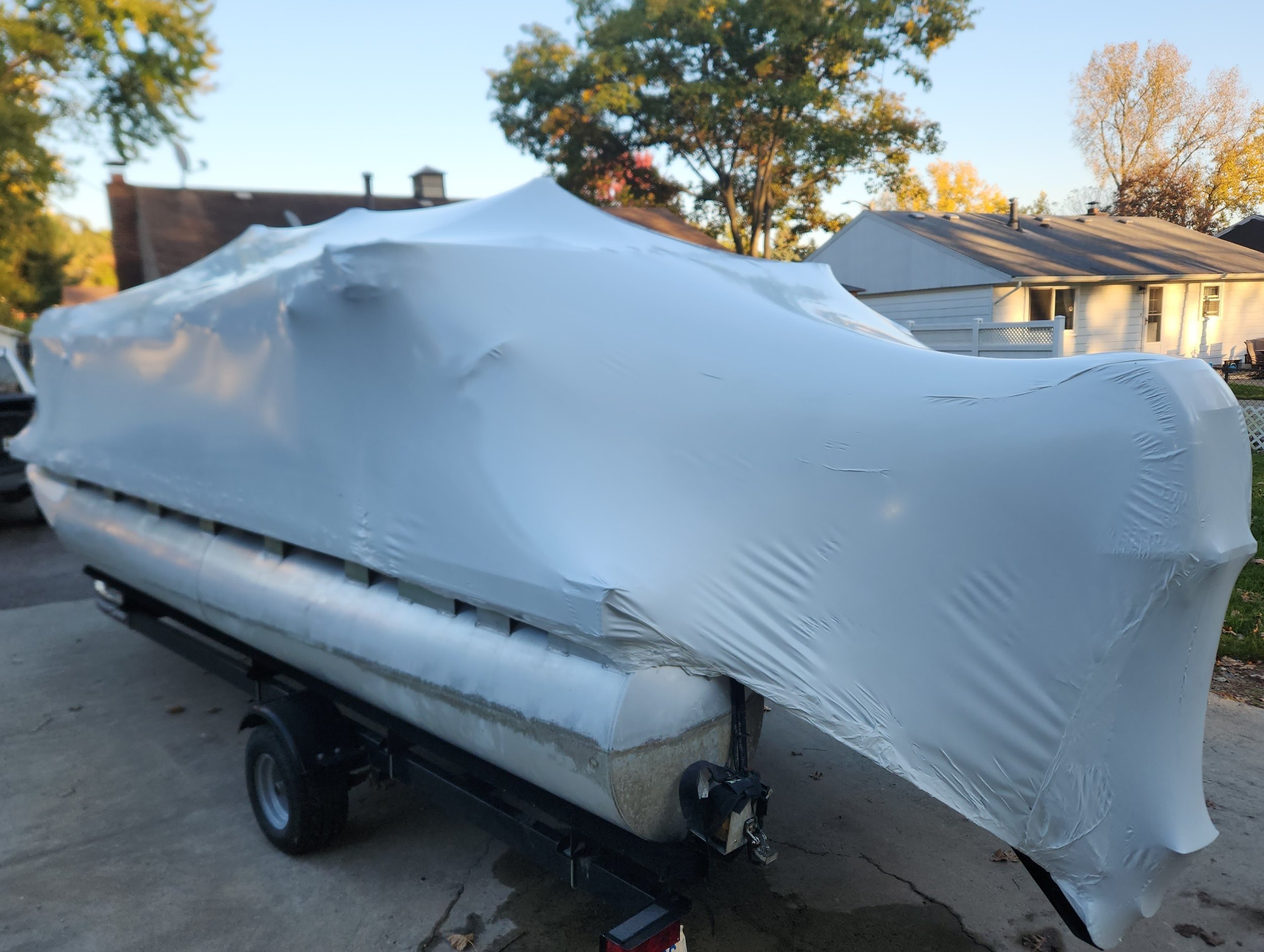 A boat covered in a white protective plastic sheet on a trailer in a driveway, with houses and trees in the background.