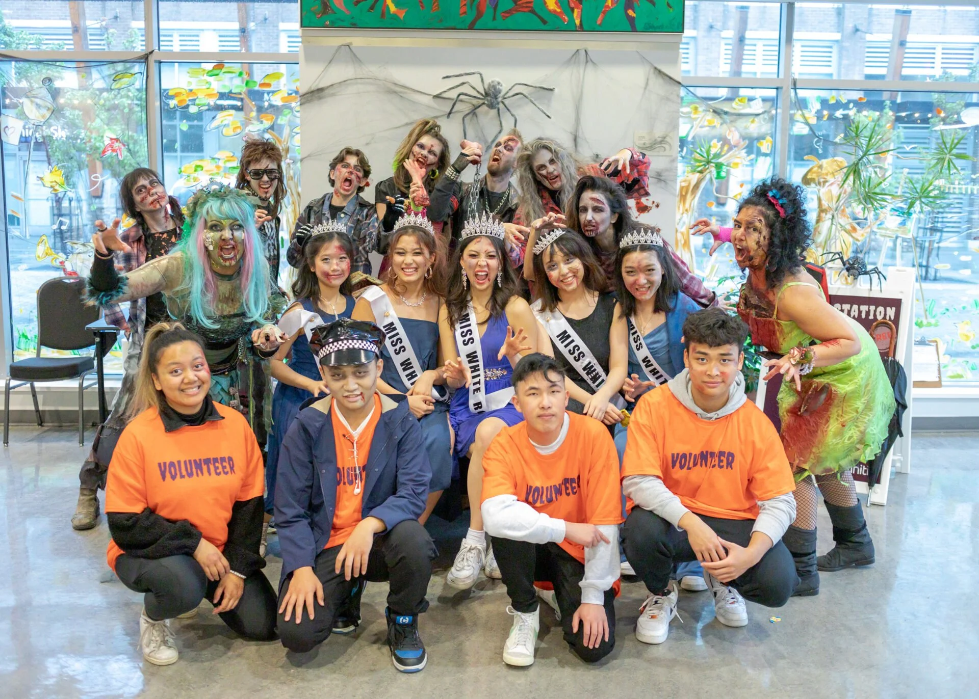 Group of young people dressed as zombies and Miss contestants, with some wearing volunteer shirts, posing together indoors near Halloween decorations, including spider web with spider, colorful artwork, and windows showing cityscapes outside.