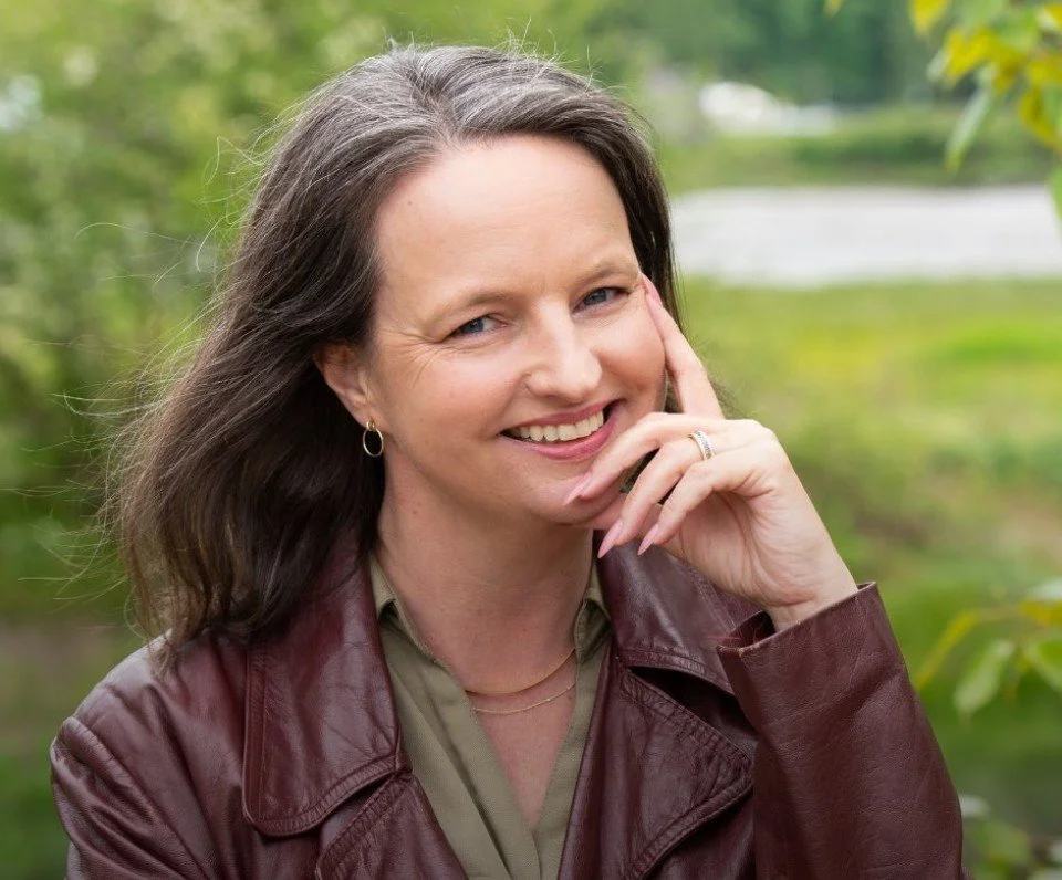 A woman with long brown hair and a smile, touching her face with her right hand, wearing a leather jacket and standing outdoors with green trees in the background.
