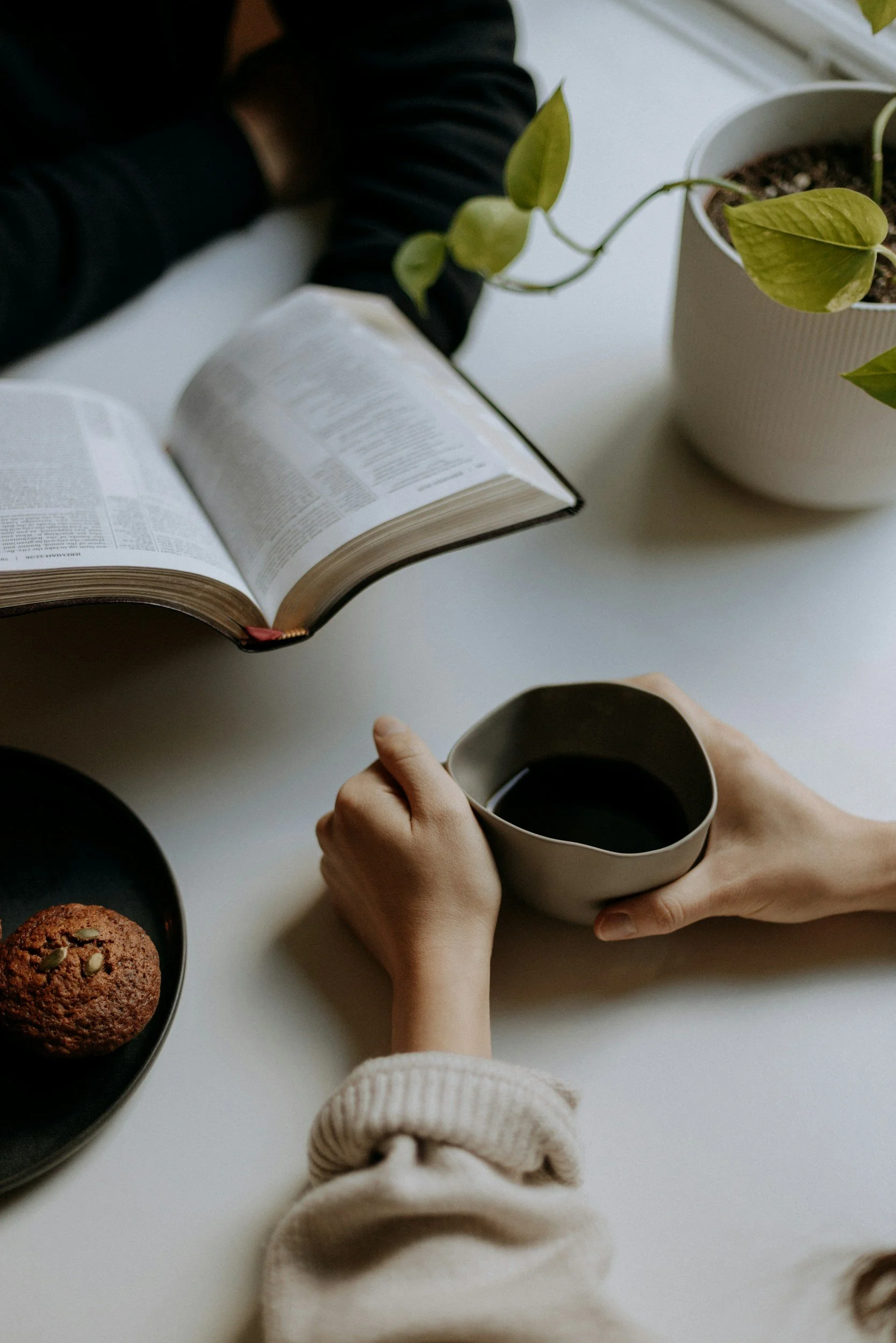 Person holding a cup of coffee, with an open book and a plate with a cookie on a white table, and a potted plant nearby.