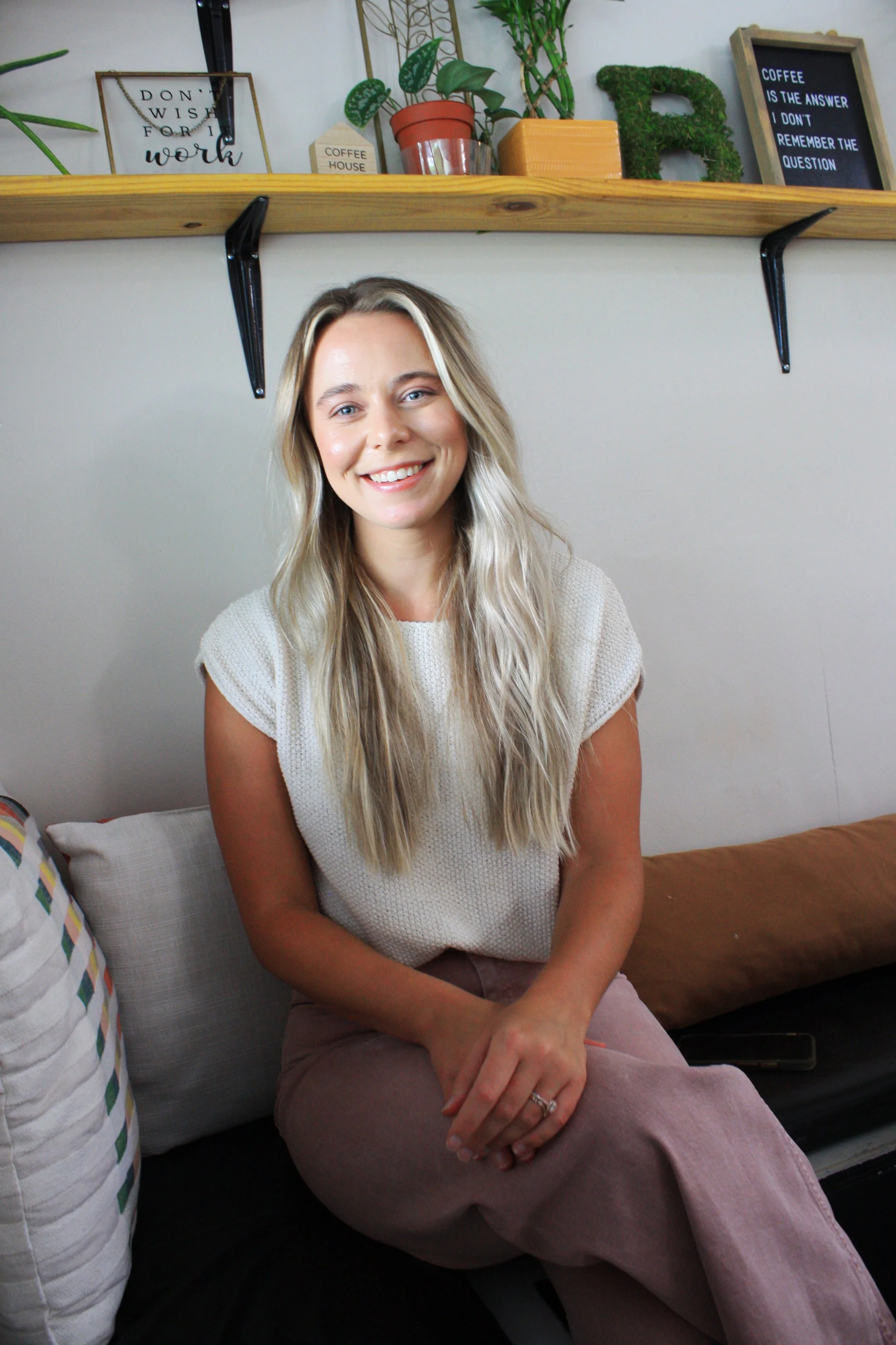 A woman with long blonde hair and a white top sitting on a black sofa, smiling at the camera in a cozy room decorated with plants and framed quotes.
