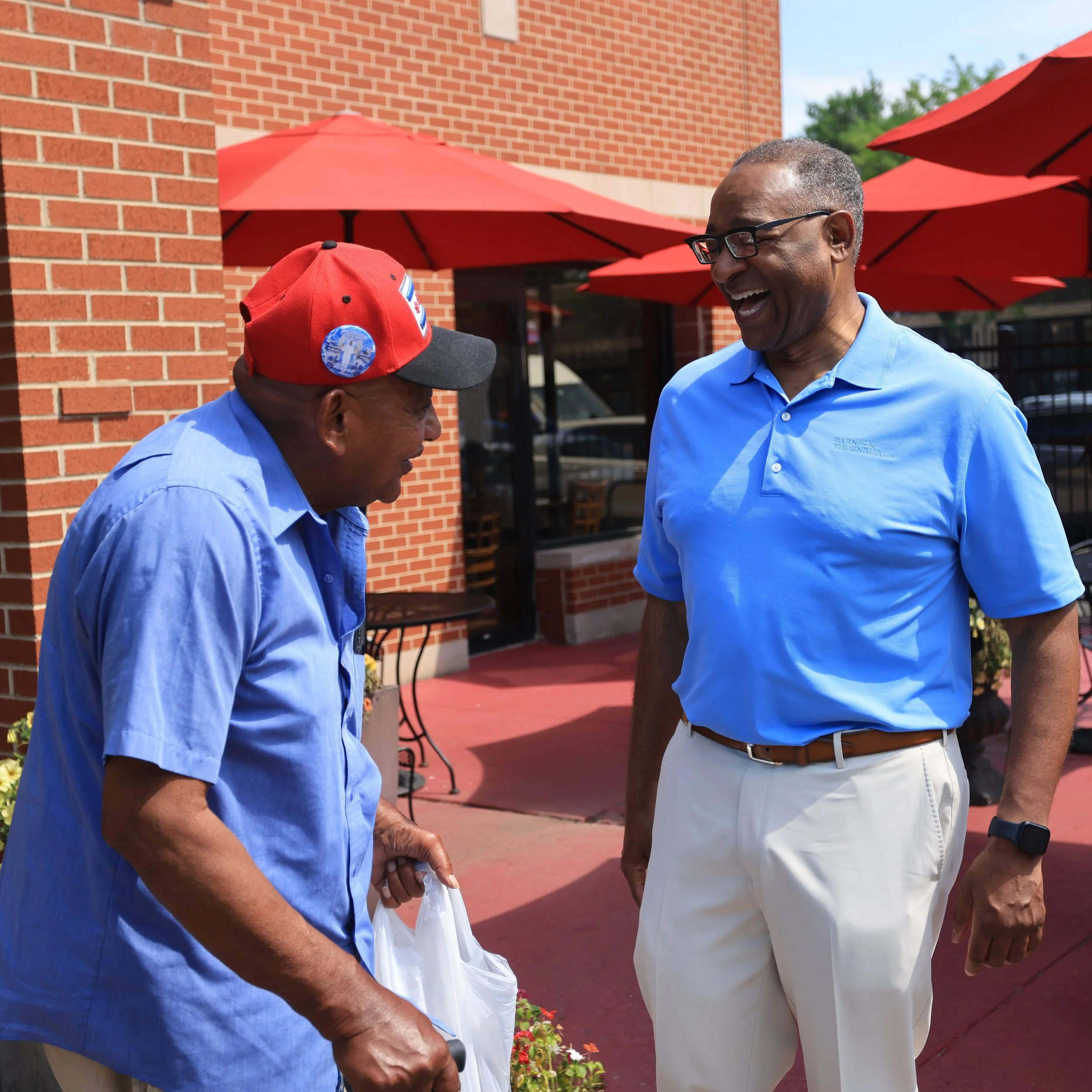 Two men smiling and chatting outdoors, one with a cane and wearing a red cap, the other is Richard Boykin. Red umbrellas shade the area, and a brick building is in the background.