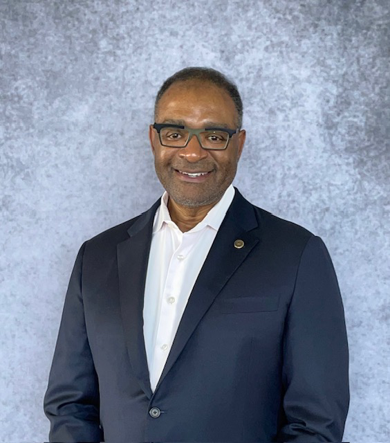 Richard Boykin in dark suit with white shirt against a gray background, smiling at the camera.
