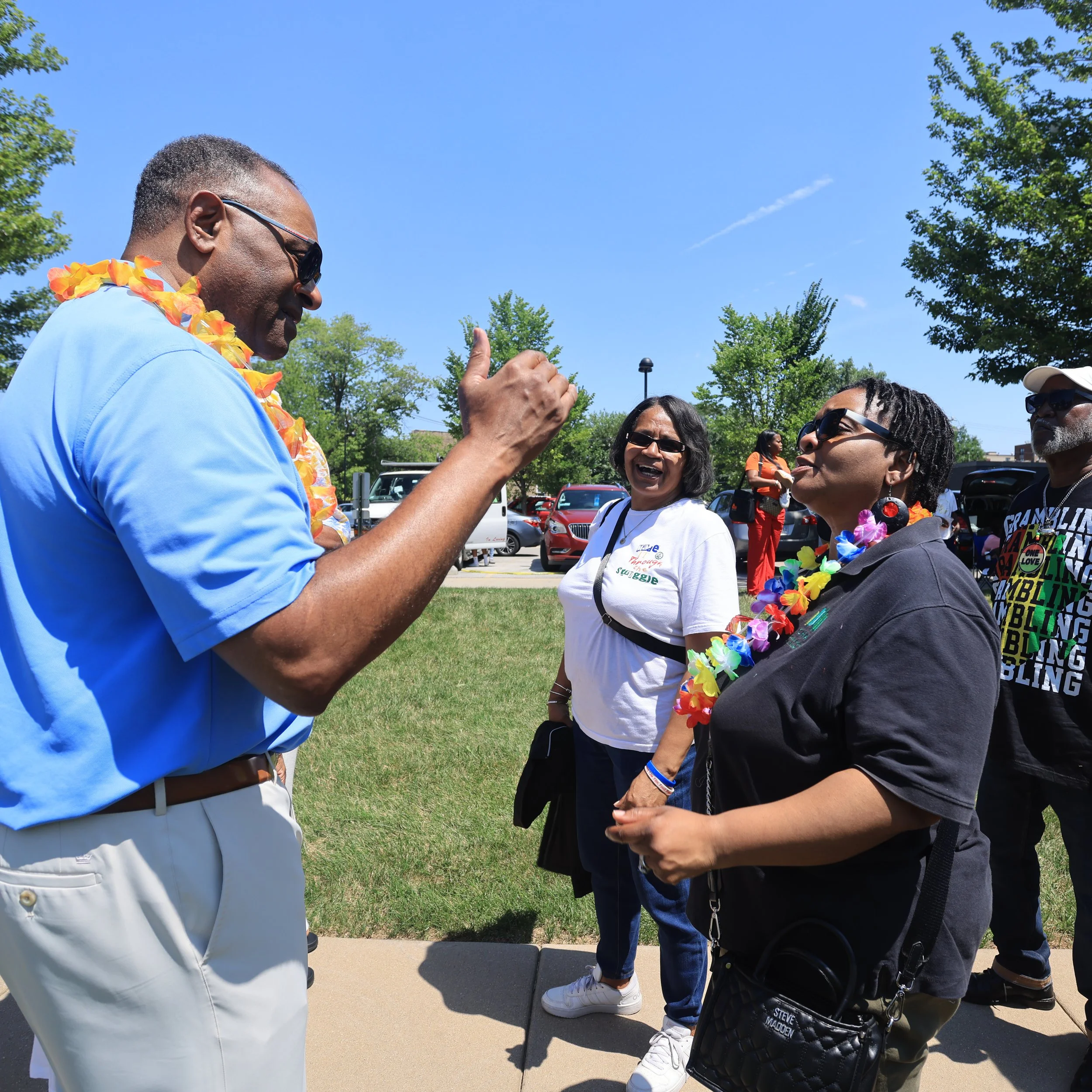 Richard Boykin talking to a group of women outdoors on a sunny day, with some of the women wearing colorful leis and laughing.